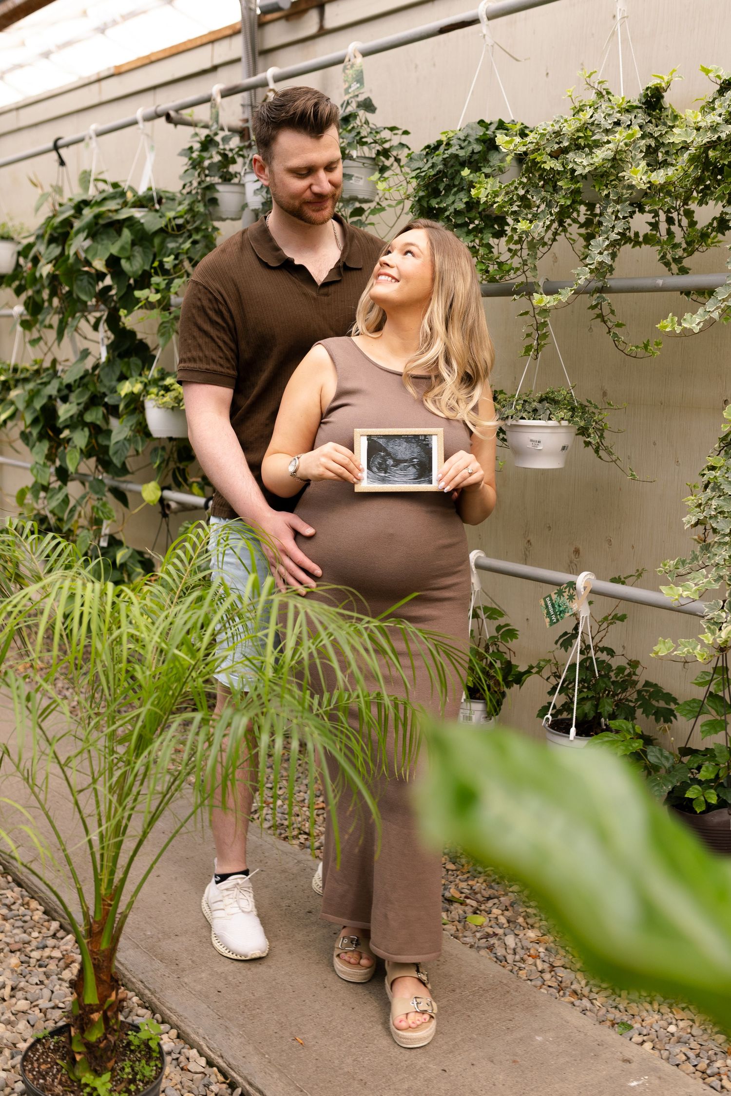 Expectant couple shares intimate moments in a plant-filled greenhouse setting.