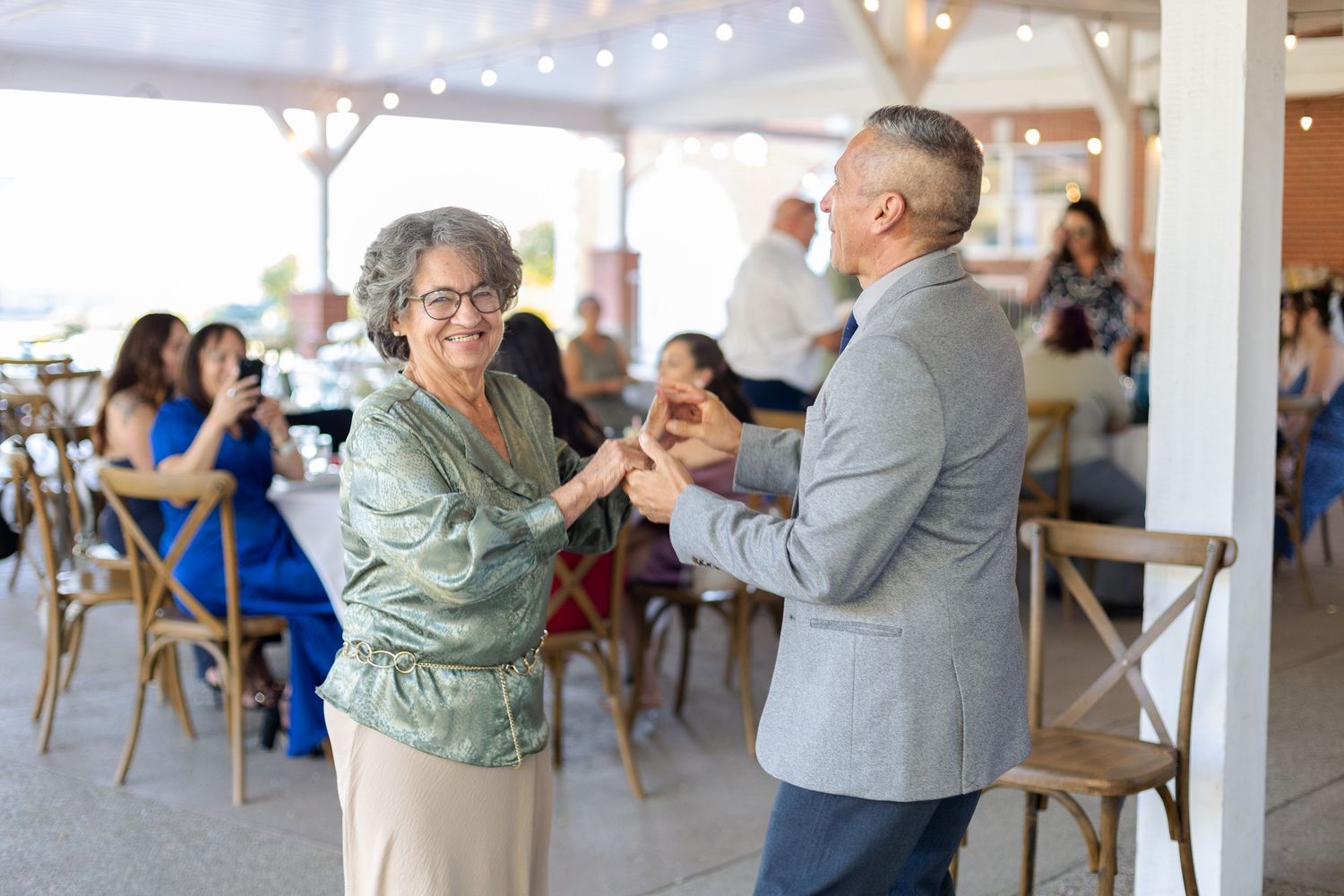 People dancing together at an outdoor reception venue decorated with string lights and wooden chairs.