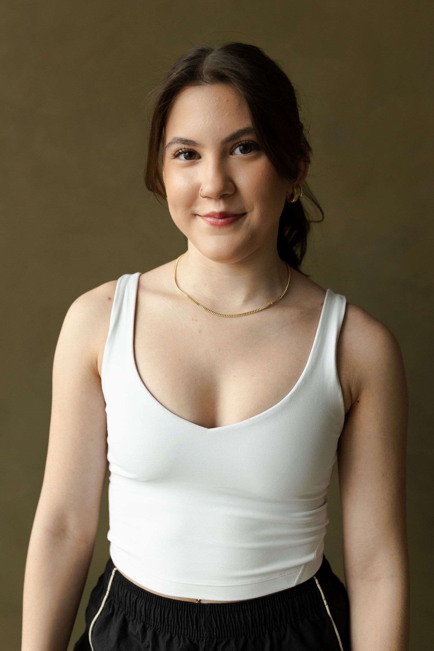 A person in a white tank top and dark pants poses against a neutral background wearing delicate jewelry.