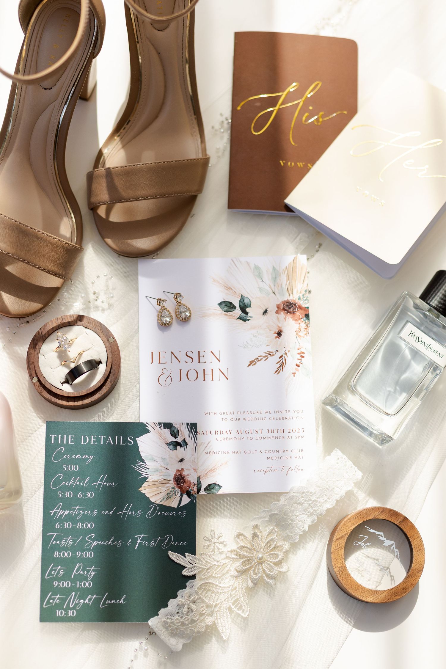 Wedding details flatlay with invitation, brown heels, perfume bottle, rings and floral decor on white background.