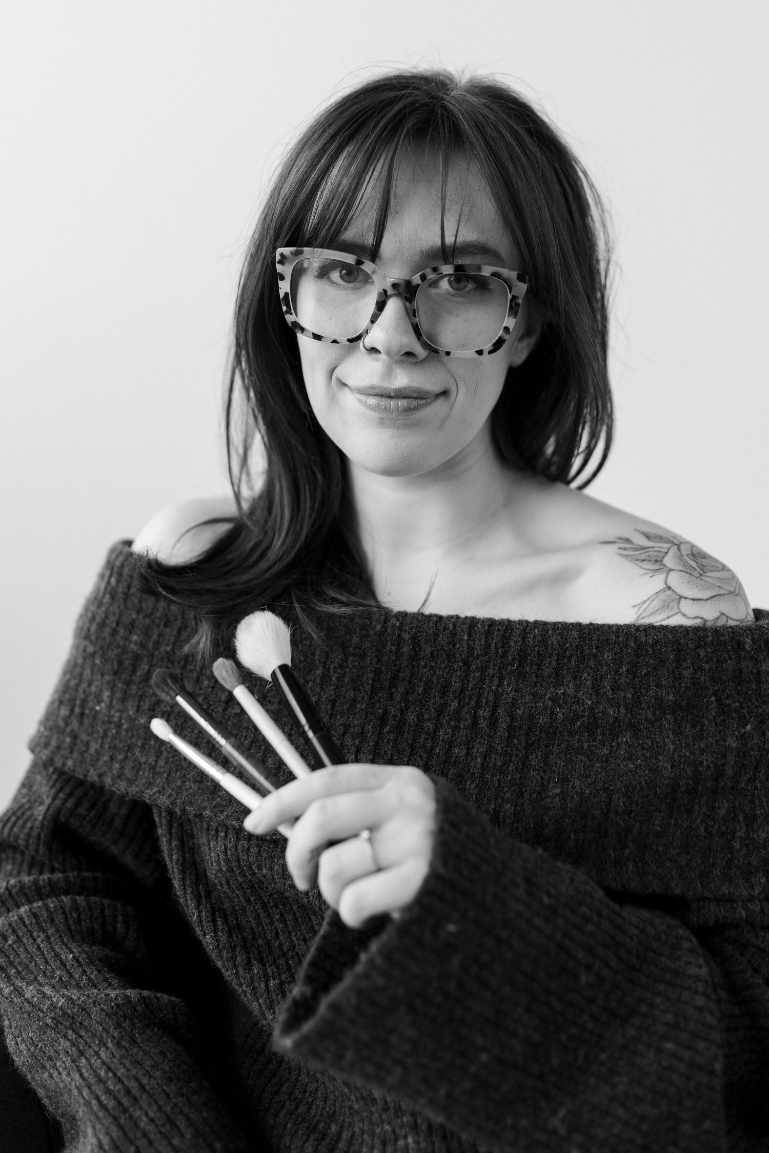 Black and white portrait of someone in glasses and off-shoulder sweater holding makeup brushes while smiling.
