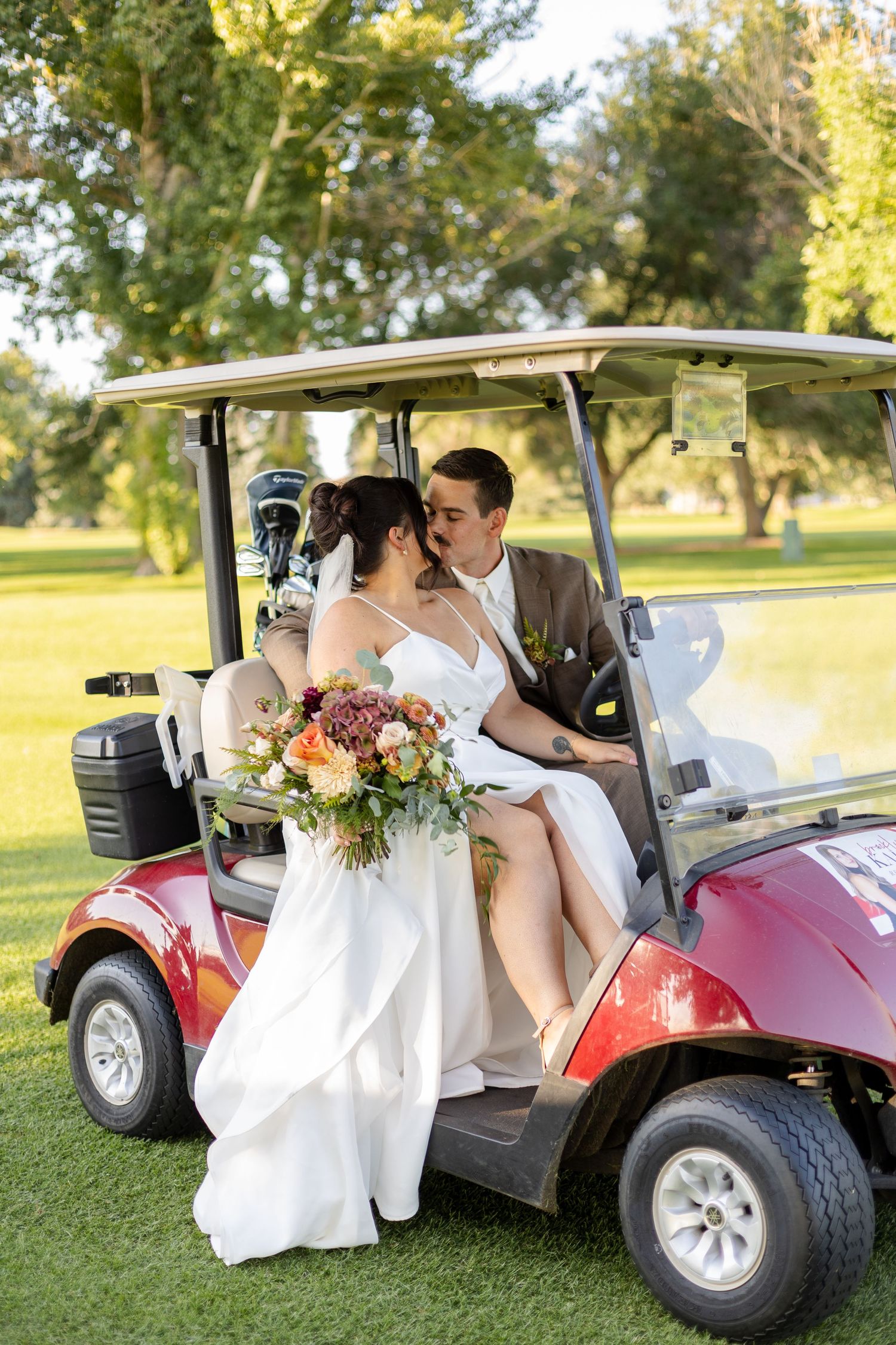 A couple shares an intimate moment in a red golf cart with a colorful floral bouquet on their wedding day.