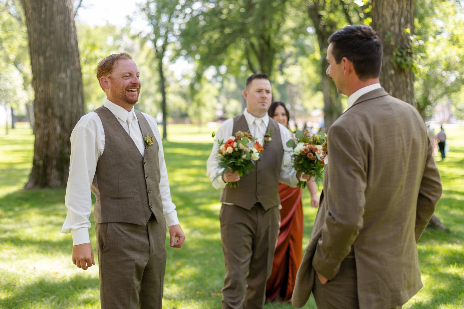 Groomsmen in brown vests share laughs while standing in a lush green park.