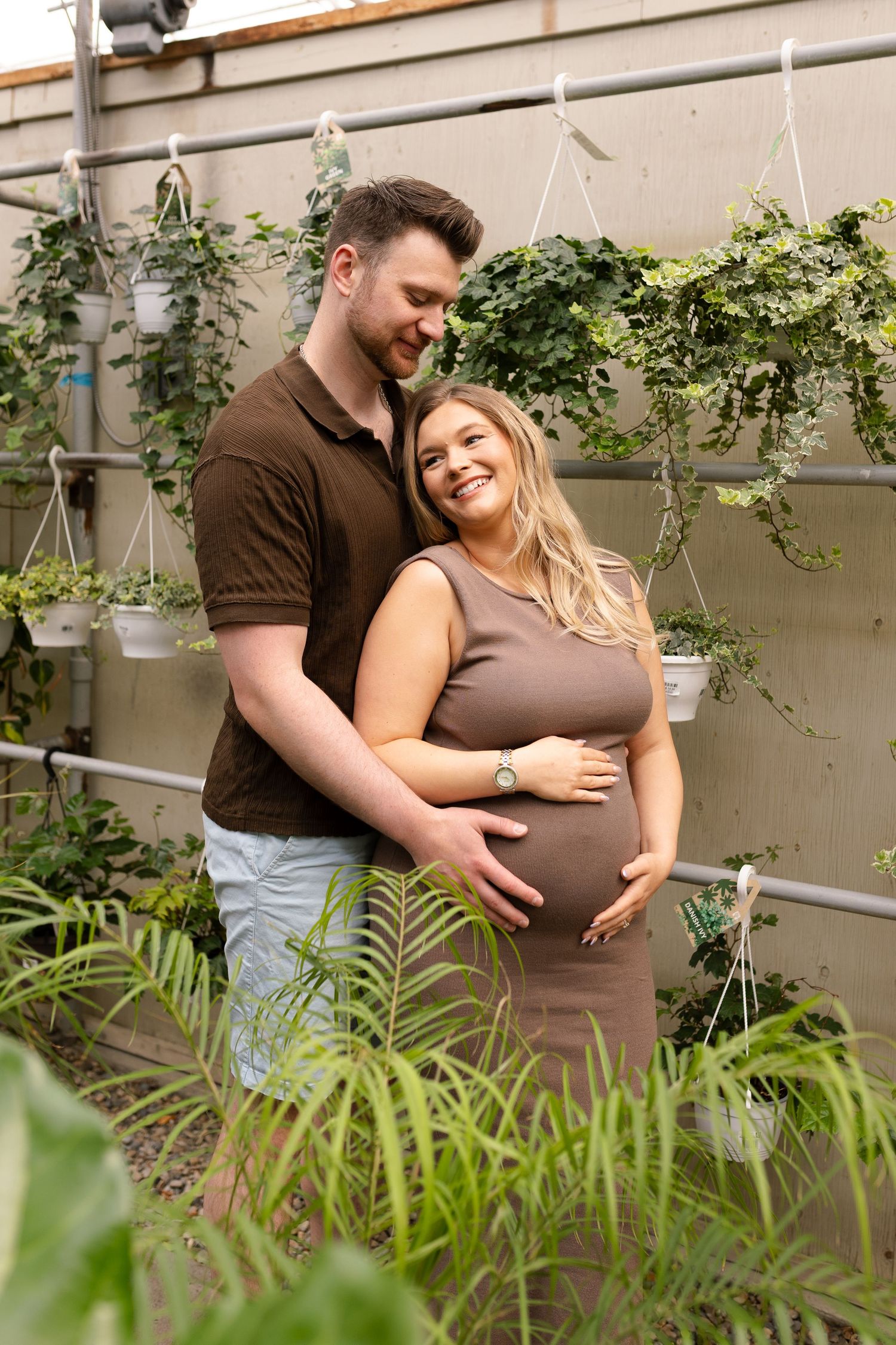 A couple embraces in a greenhouse setting surrounded by hanging plants and greenery.