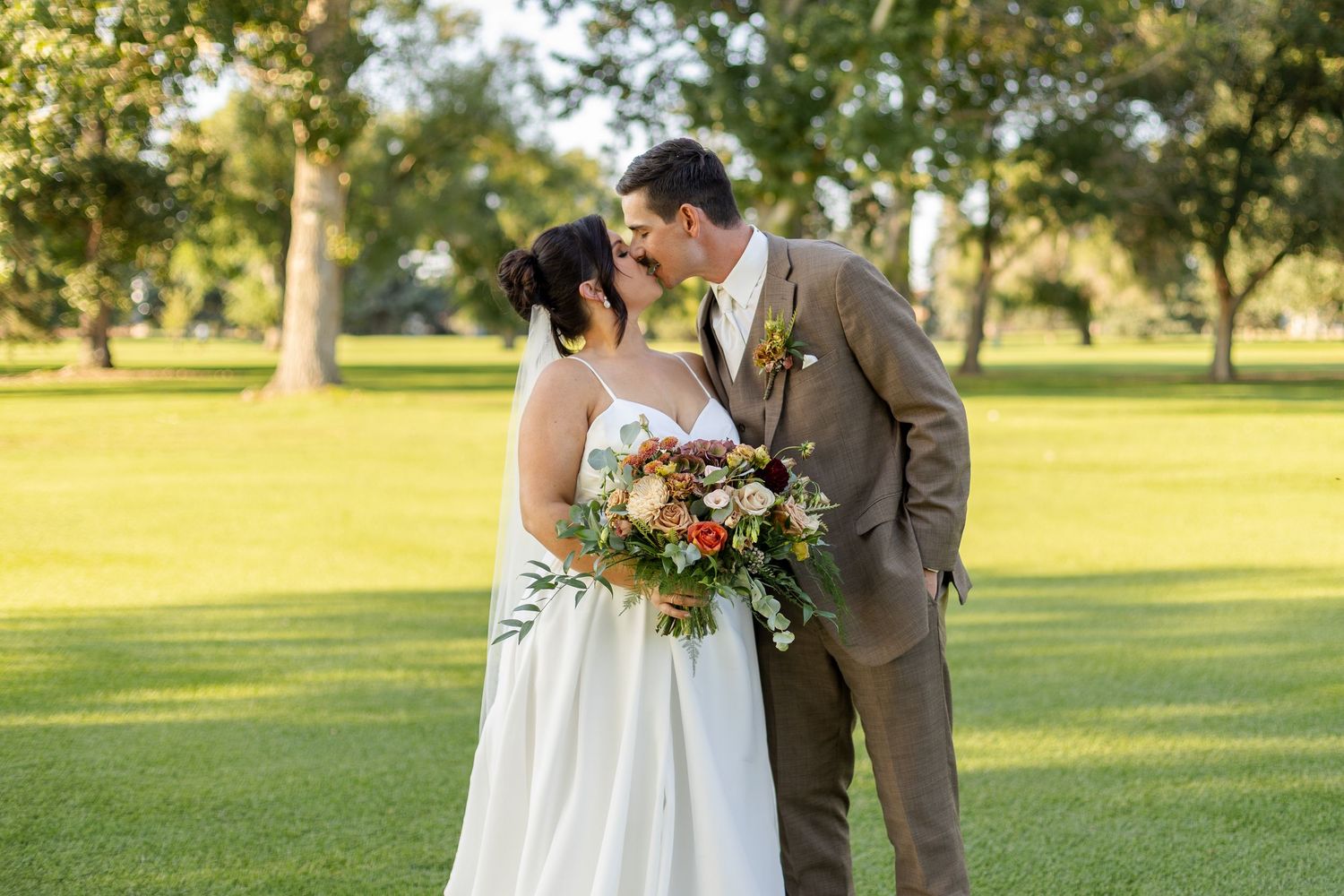Wedding couple poses in formal attire on sunny grass field with trees in background.