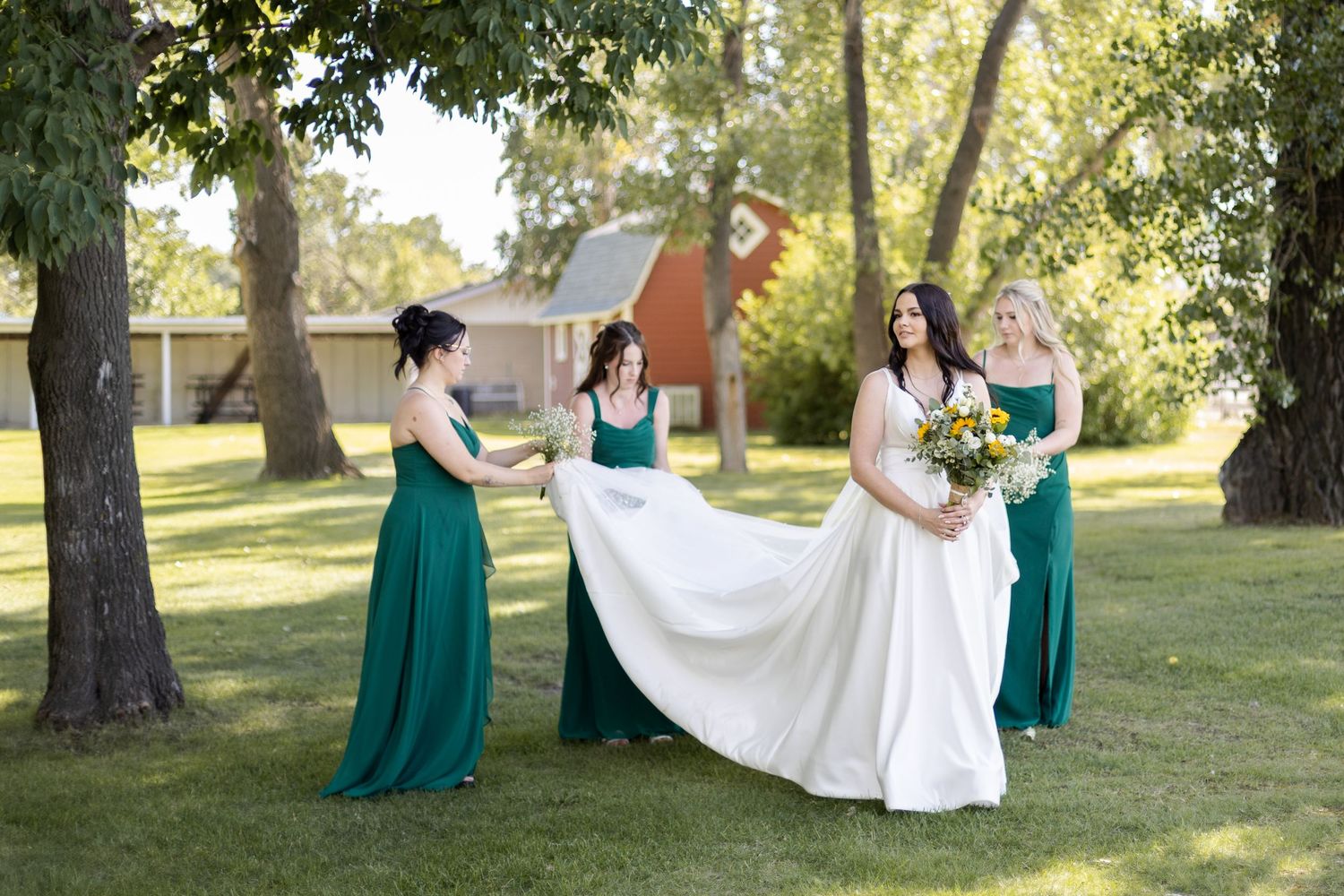 Bridesmaids in emerald green dresses help arrange a flowing white wedding dress train on sunny lawn.