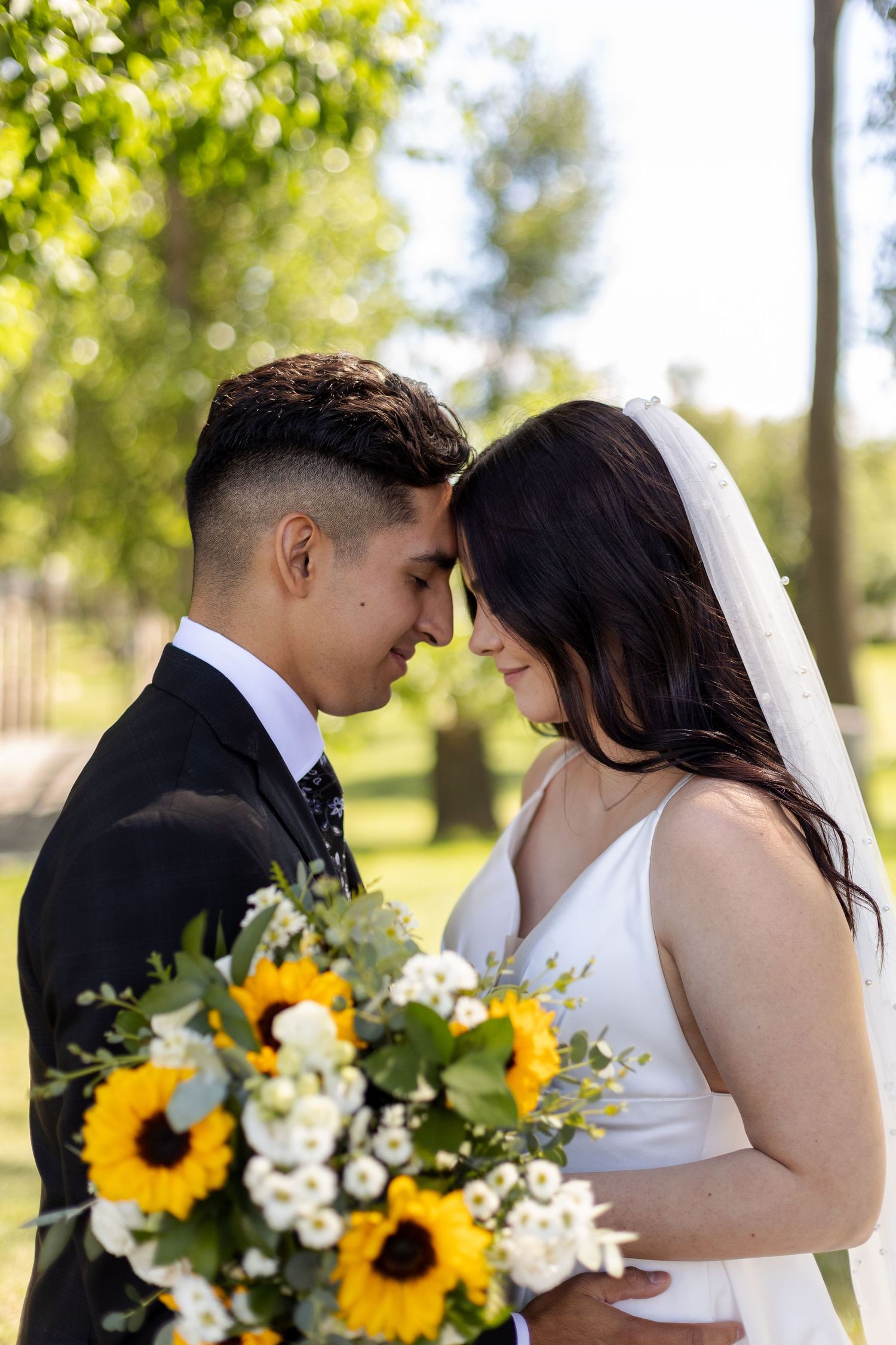 Romantic wedding couple poses in sunlit garden with bright sunflower and white floral bouquet.