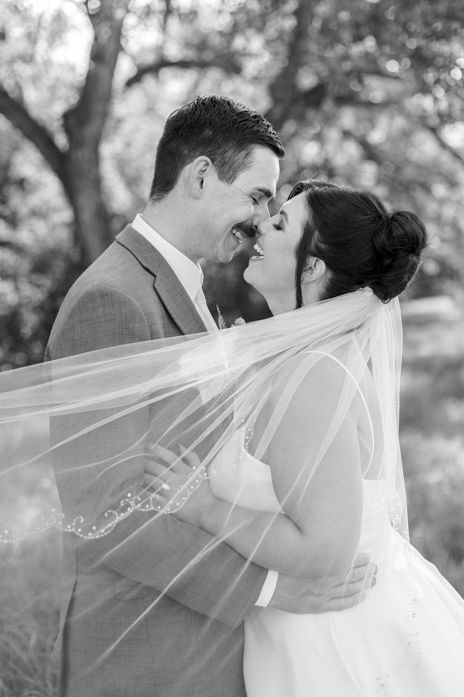 Wedding couple shares romantic kisses through flowing white veil in black and white outdoor portrait series.