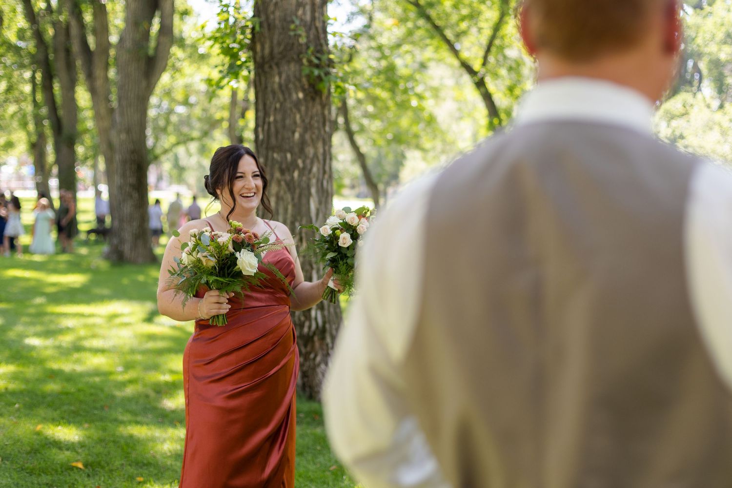 Wedding party in burgundy dresses and tan suits poses outdoors in a sunlit garden setting.