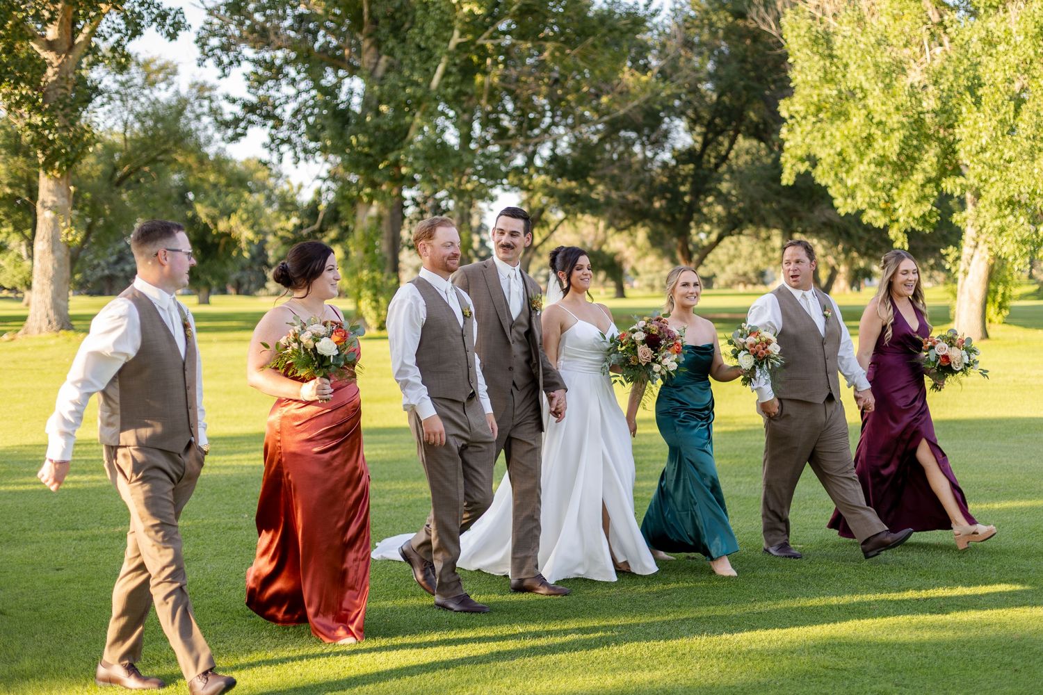 Wedding party members walk and pose together on sunny green lawn.