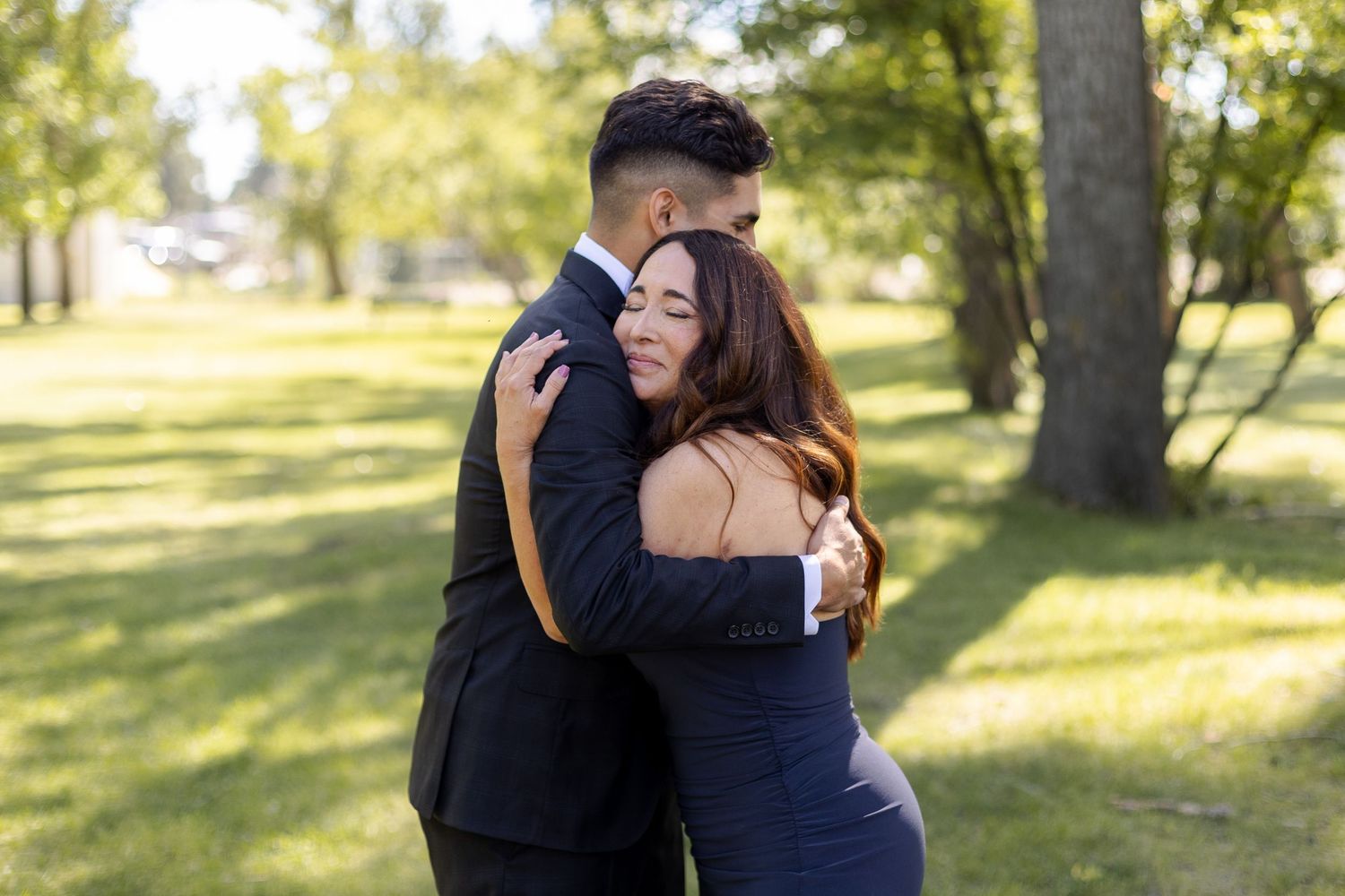 A couple embraces outdoors in a sunlit park setting with trees and golden light in the background.