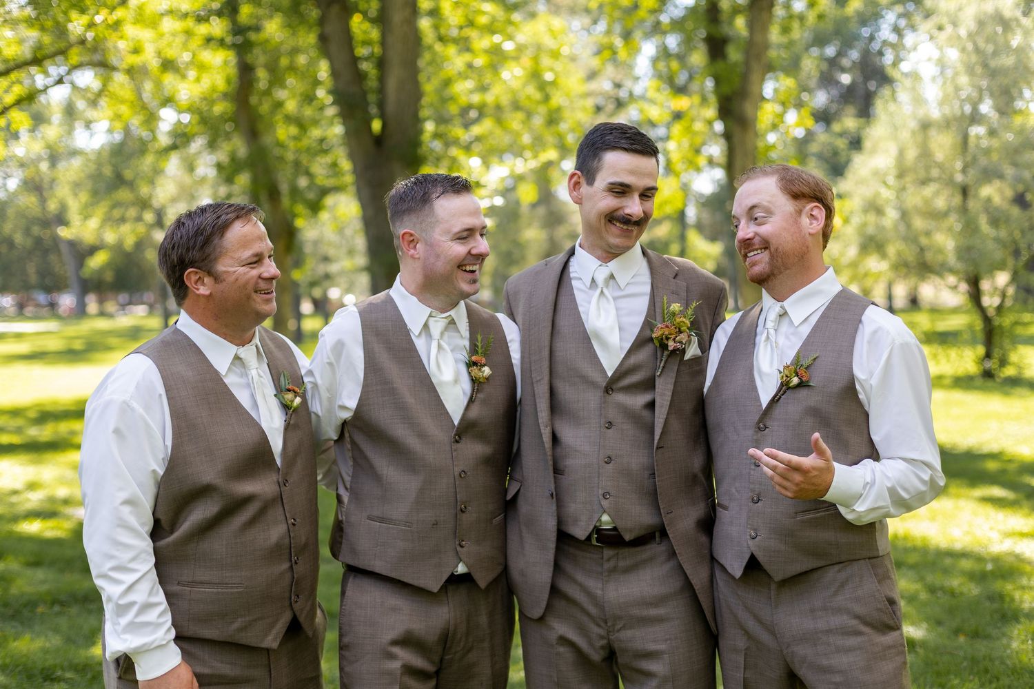 Groomsmen in brown vests and white shirts gather for candid moments in a sunny outdoor setting.