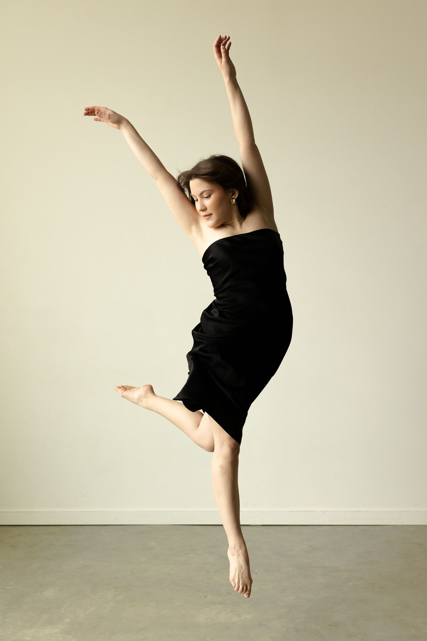 Graceful dance pose with arms raised in black dress against neutral wall.