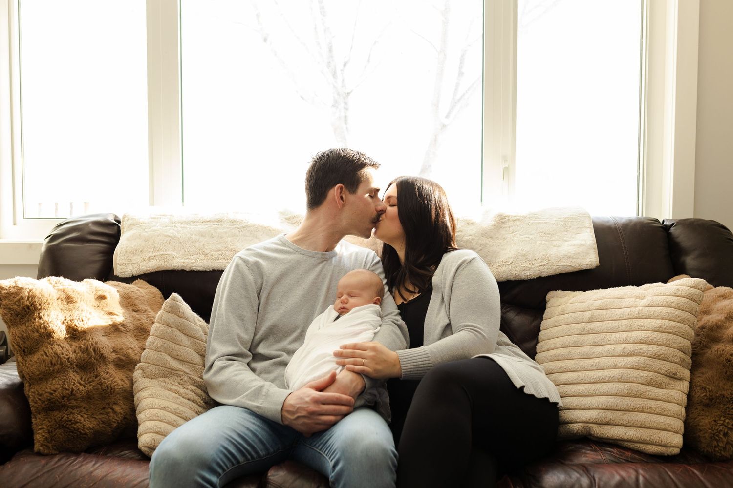 Family shares intimate moment on couch with newborn baby surrounded by decorative pillows.