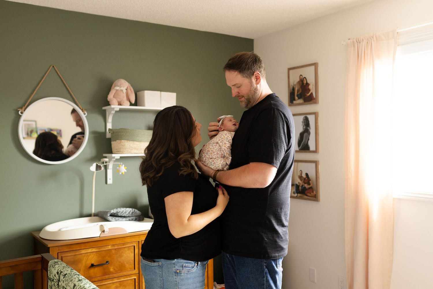 A couple stands together in a nursery room with sage green walls and a wooden dresser.