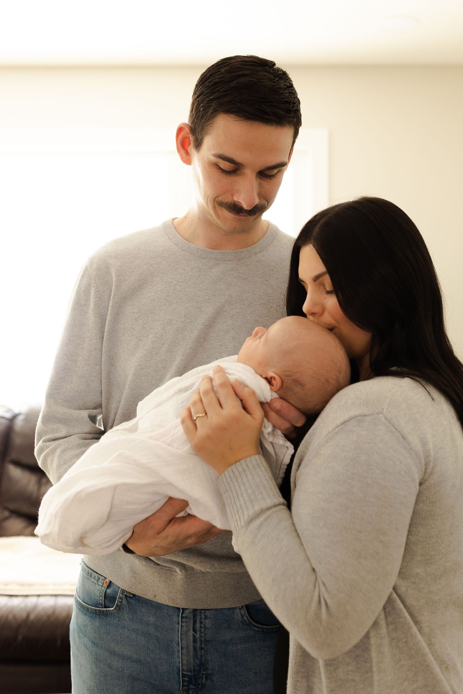 Parents embrace while holding their sleeping newborn baby dressed in white.