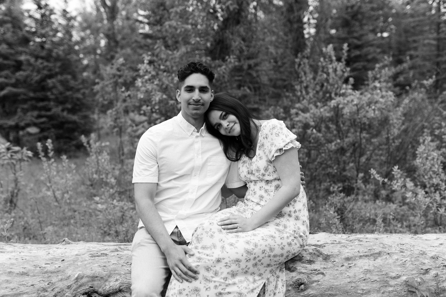 A black and white portrait of a couple sitting together on a fallen log in nature.