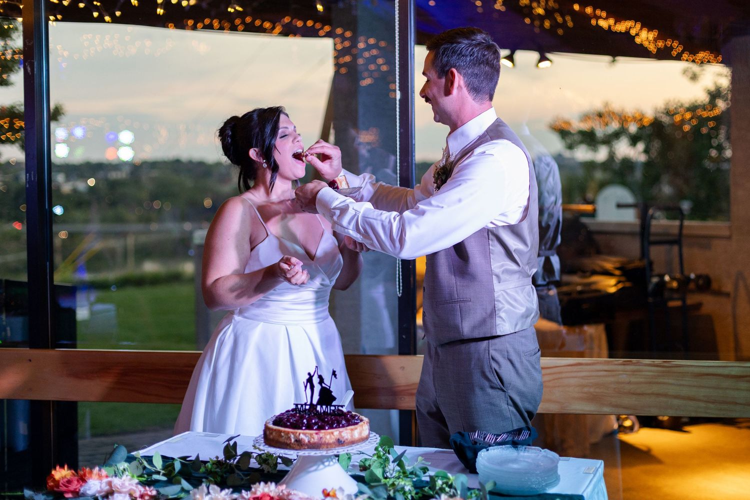 Newlyweds share intimate moment while cutting cake at sunset reception with string lights overhead.