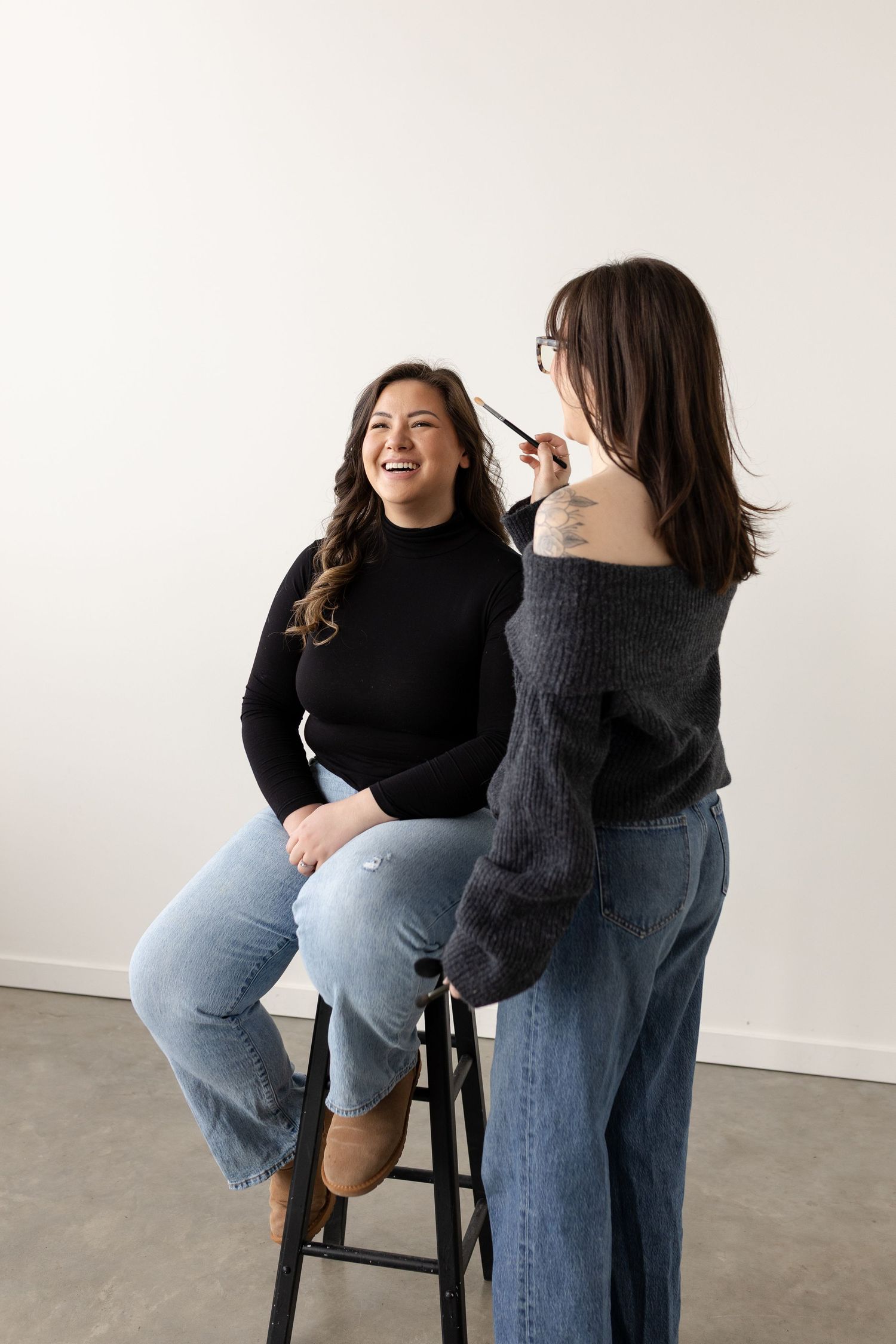 Two people in black sweaters and jeans share joyful laughter during a casual photoshoot against a white background.