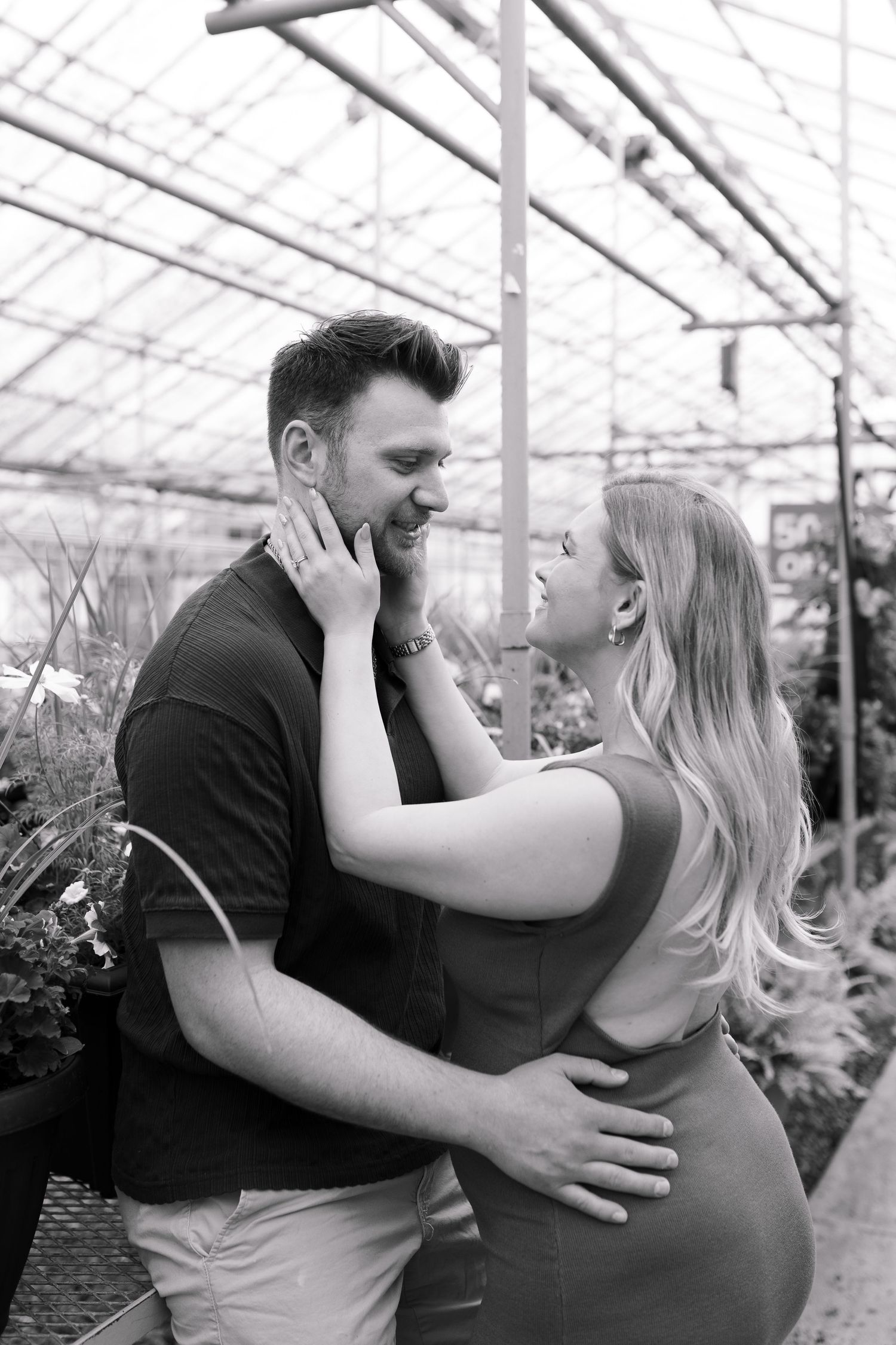 Black and white maternity photo capturing intimate moment between couple in greenhouse setting.