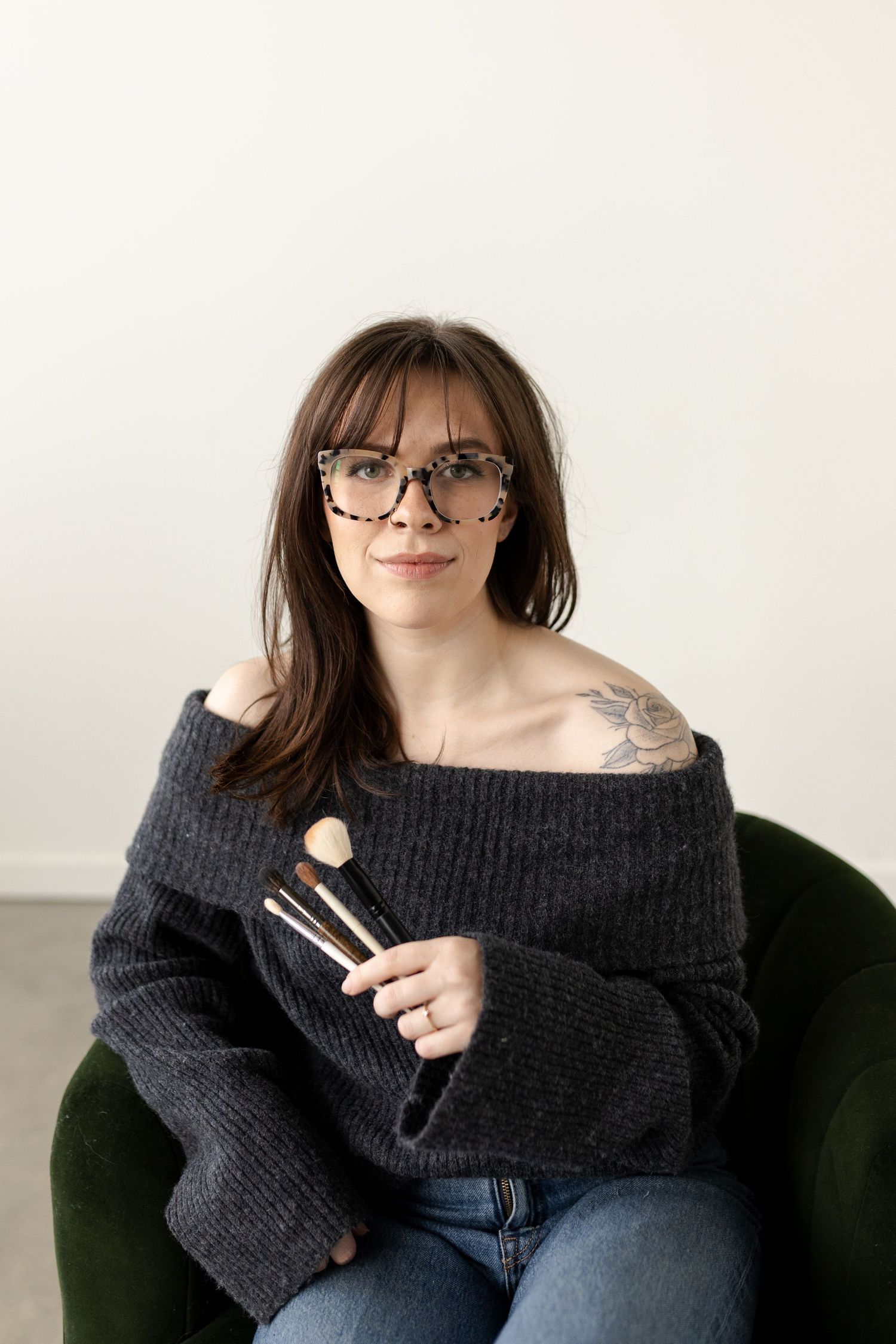 Person in tortoise shell glasses and off-shoulder sweater holds makeup brushes while sitting in green velvet chair.