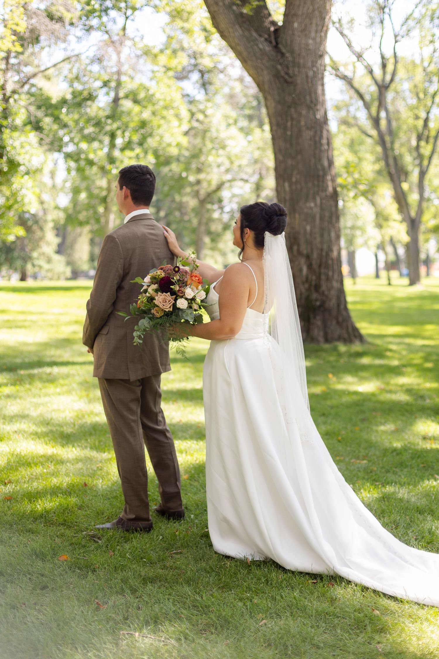 A wedding couple shares a romantic first look moment in a sunlit park with tall trees.