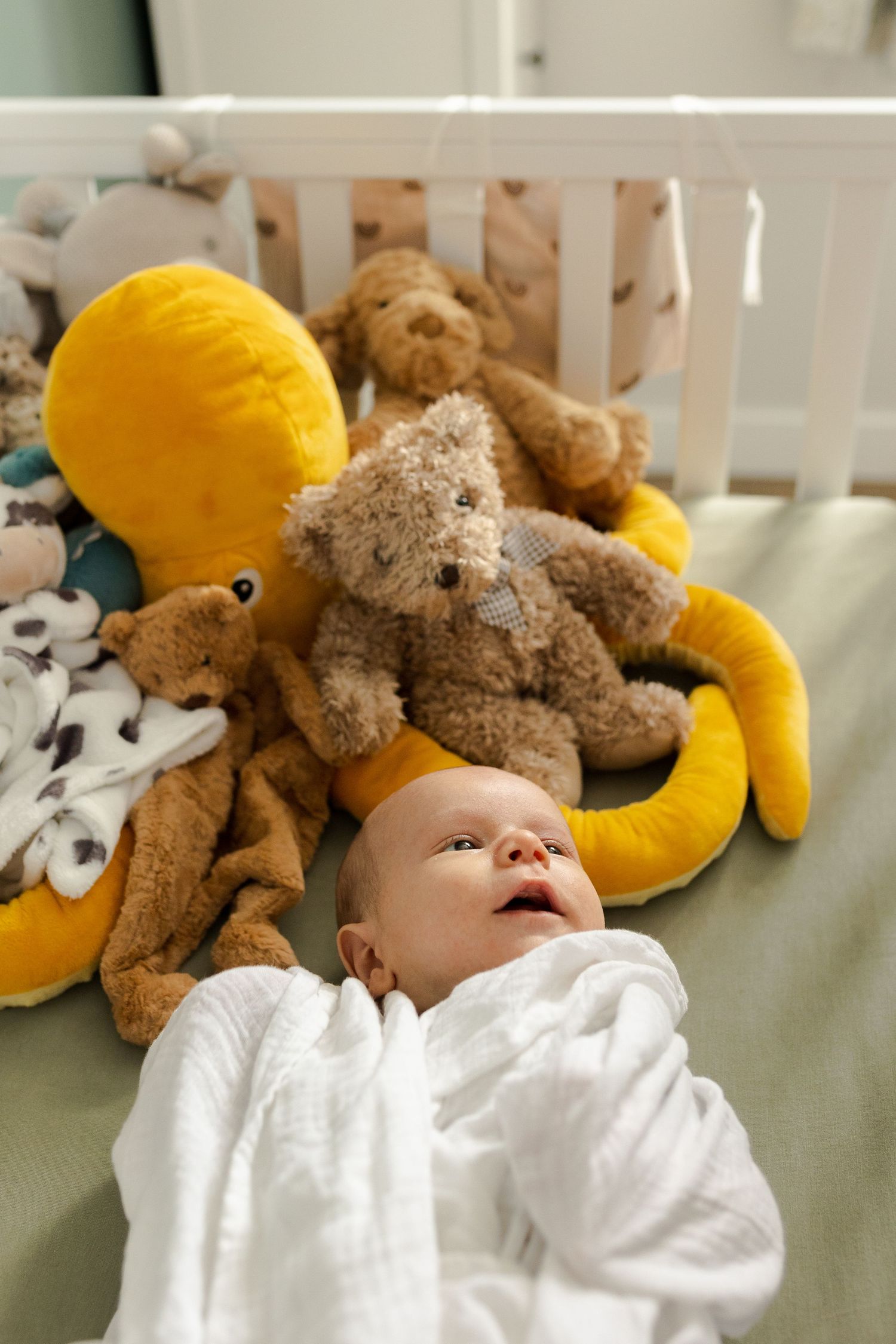 Infant sleeping peacefully in white bedding surrounded by plush teddy bears and yellow toys.