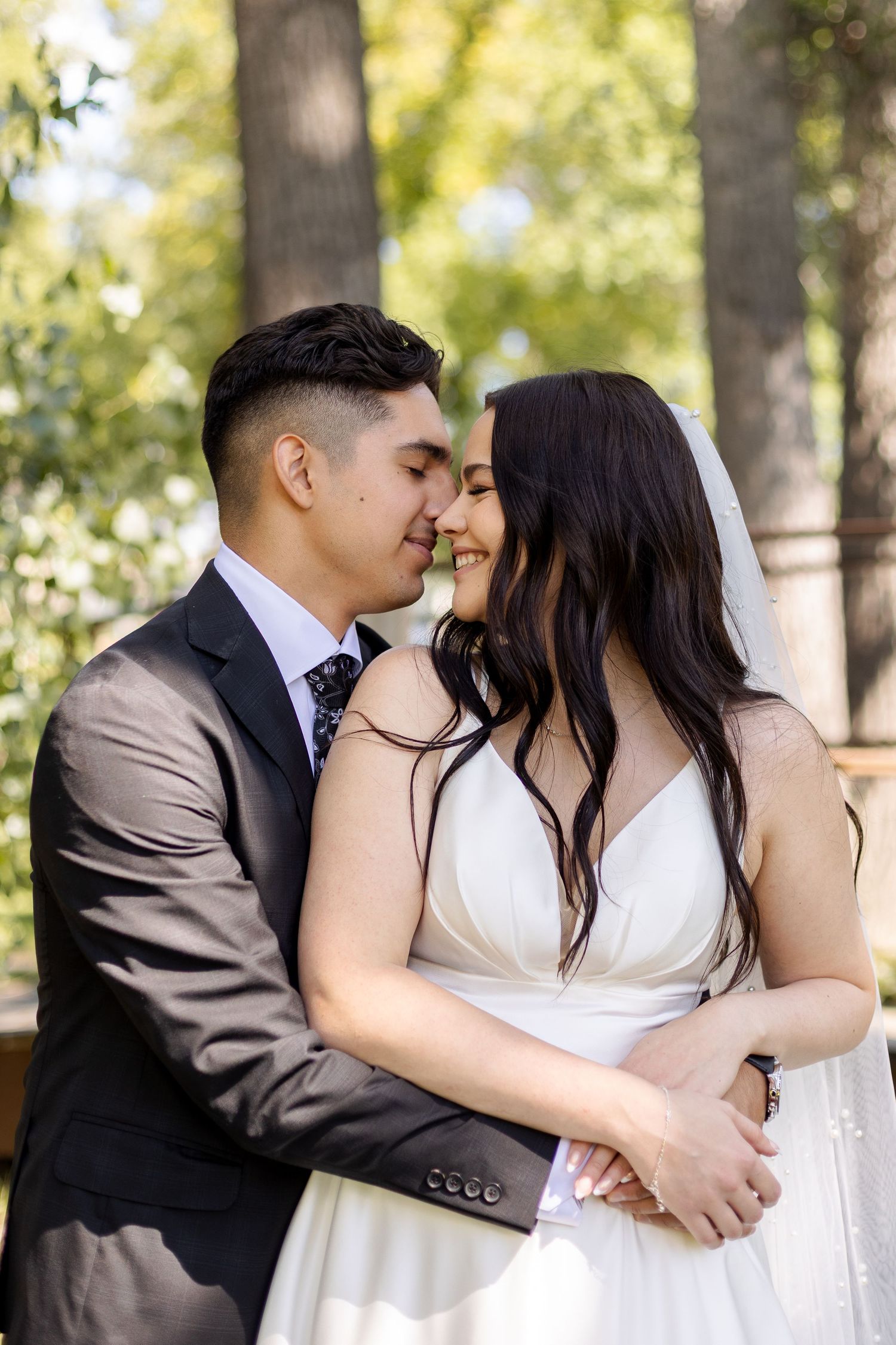 Intimate wedding portrait of newlyweds embracing in a wooded setting with sunlit trees in the background.