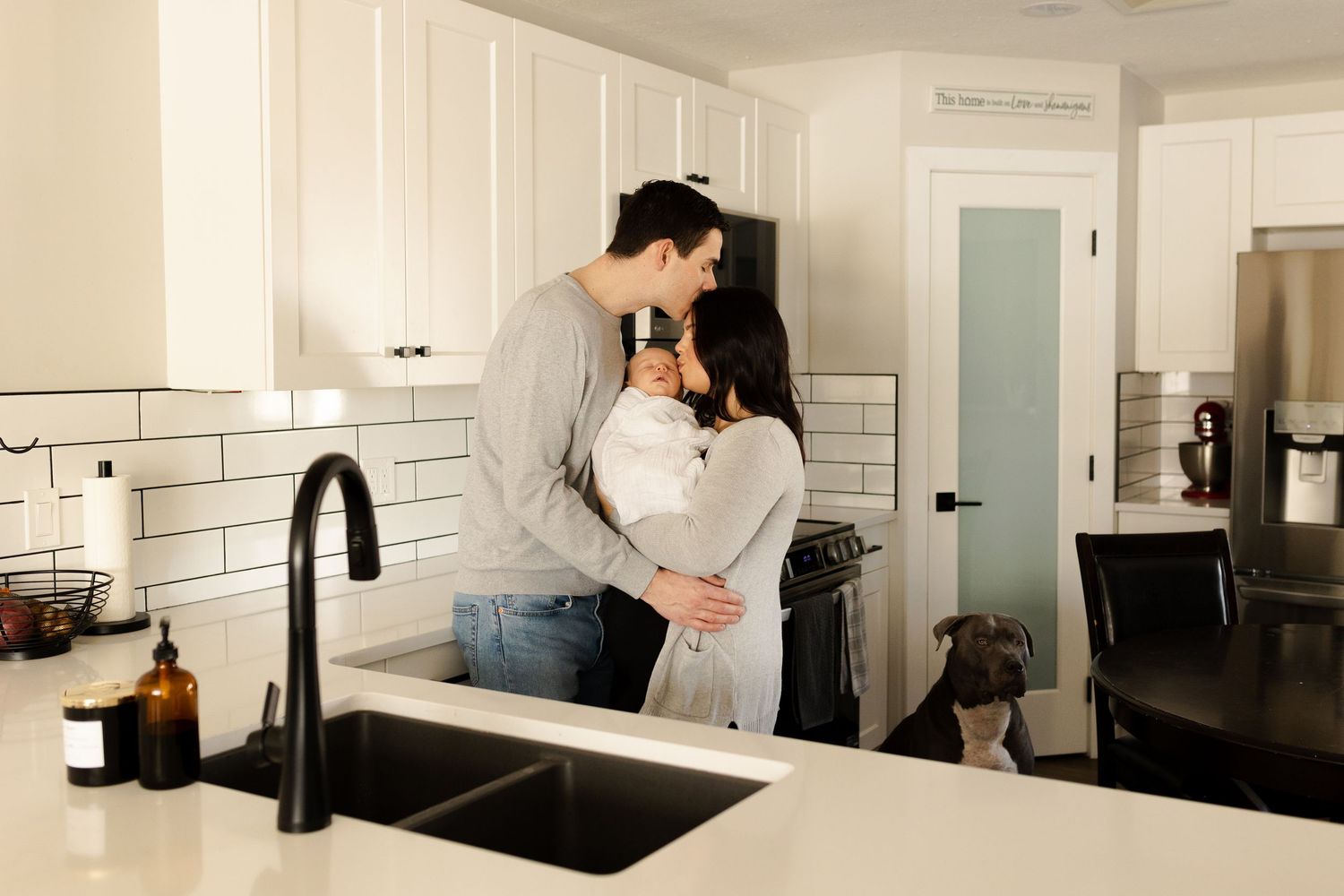 A romantic couple embraces in a bright modern kitchen with white subway tile walls and dark countertops.
