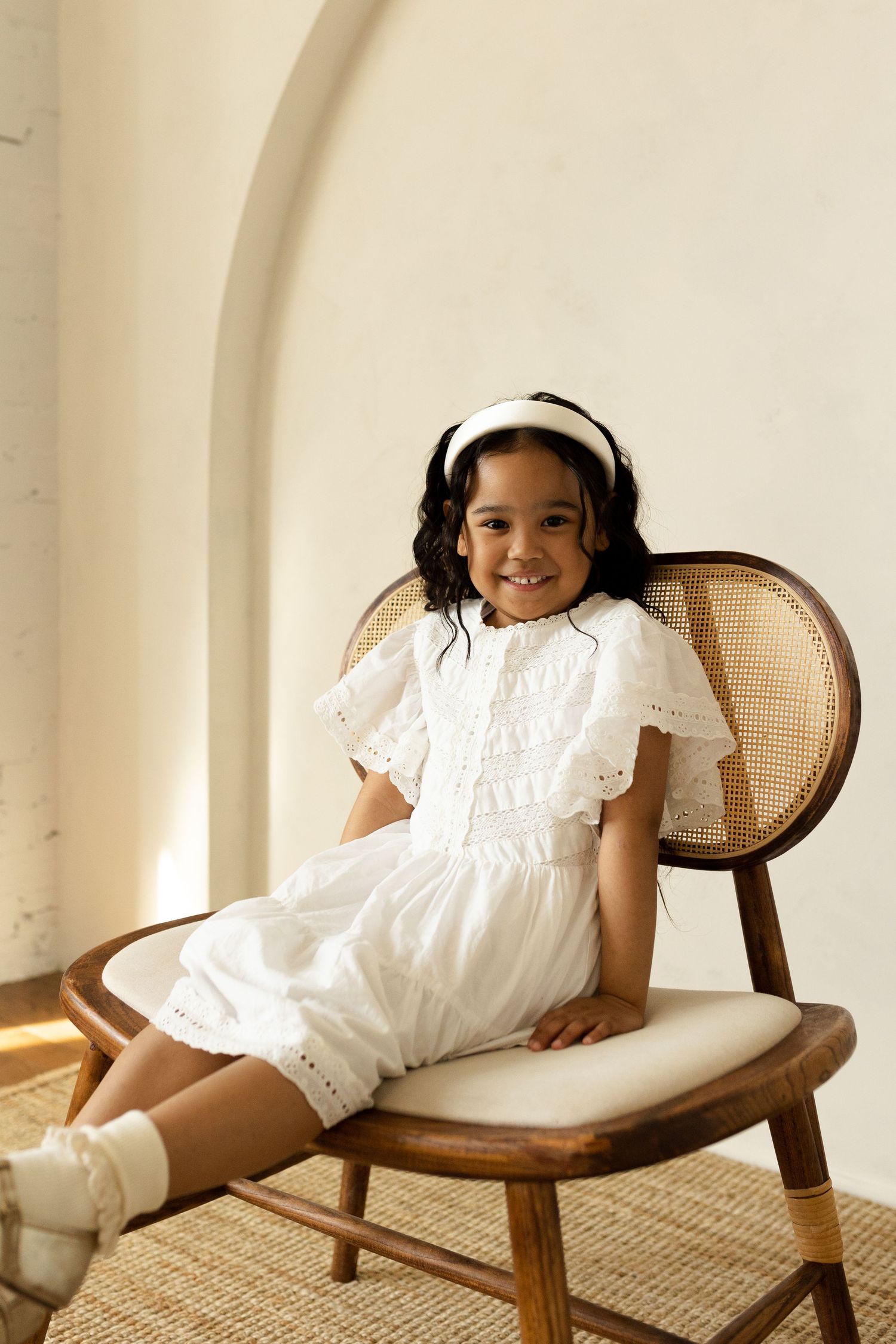 Studio portraits on a vintage cane chair showing playful poses in white summer dresses.