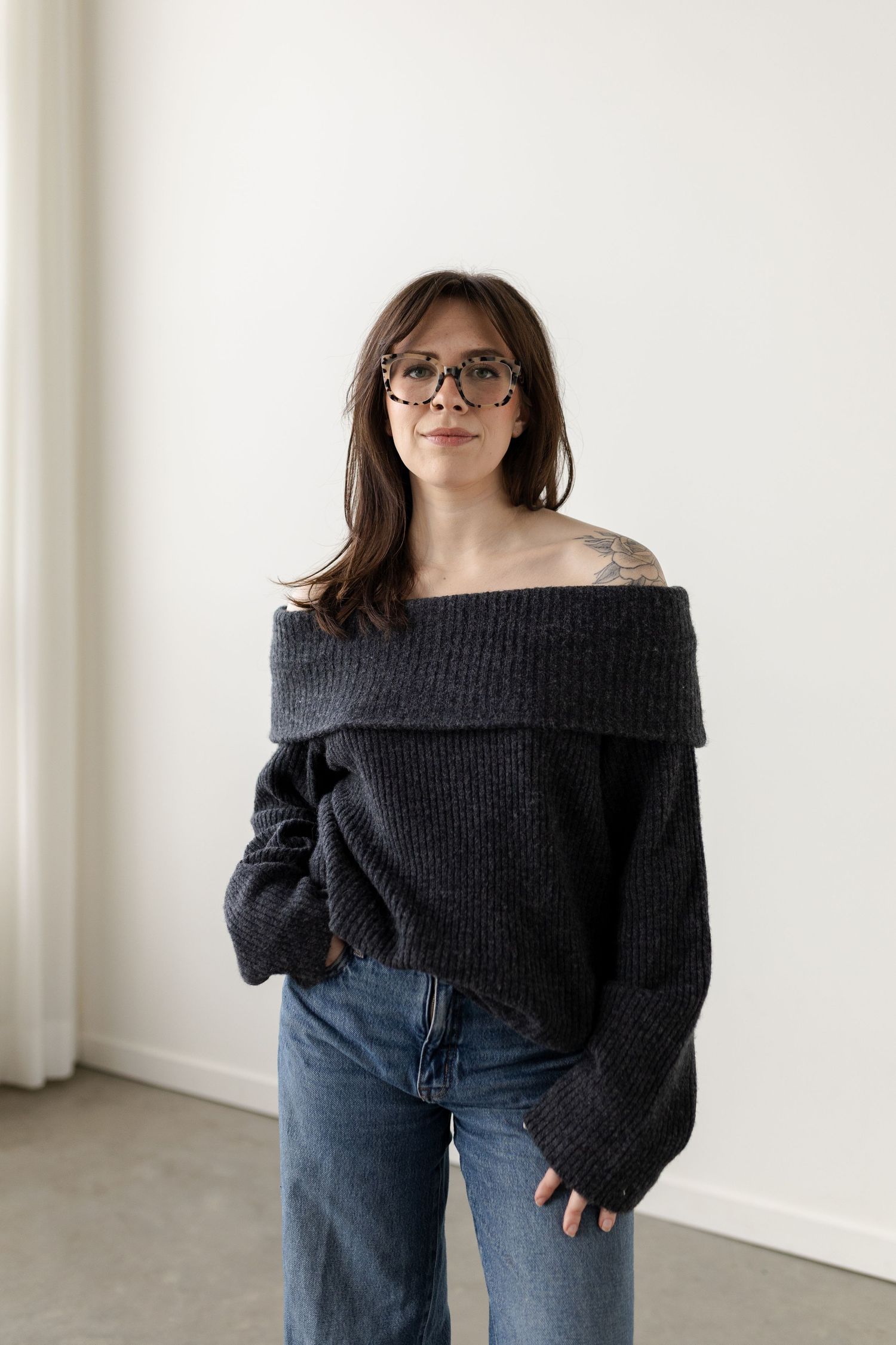 Casual outfit featuring an off-shoulder black knit sweater paired with blue jeans in a minimalist white room setting.
