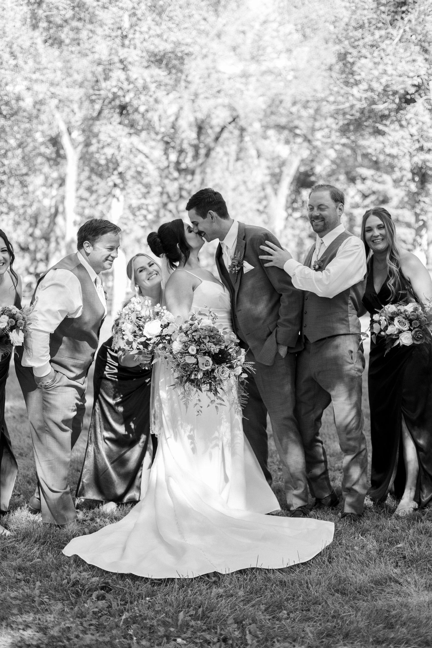 Black and white photo of wedding party laughing together on grassy lawn.