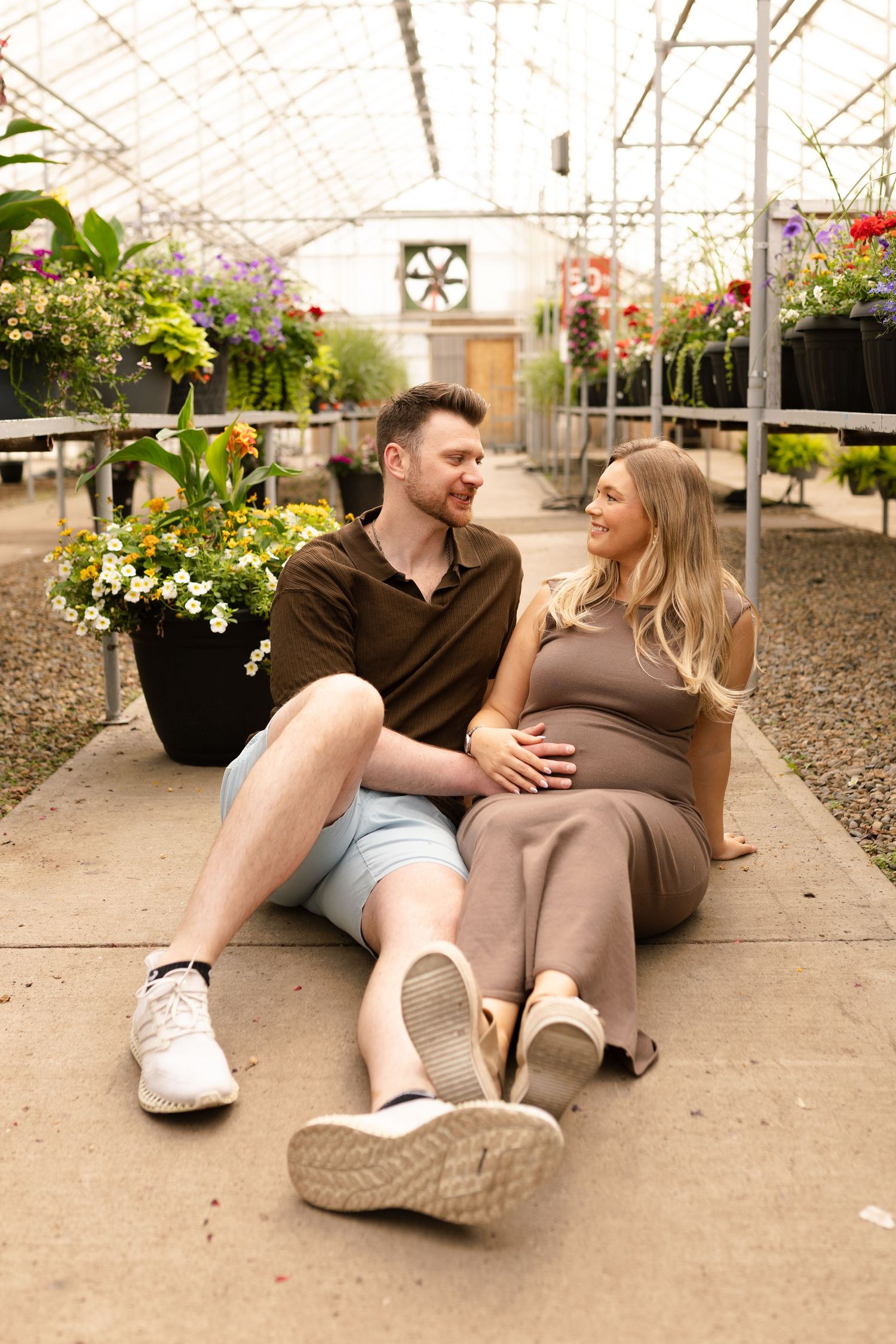 Couple relaxing on greenhouse floor surrounded by flowering plants.