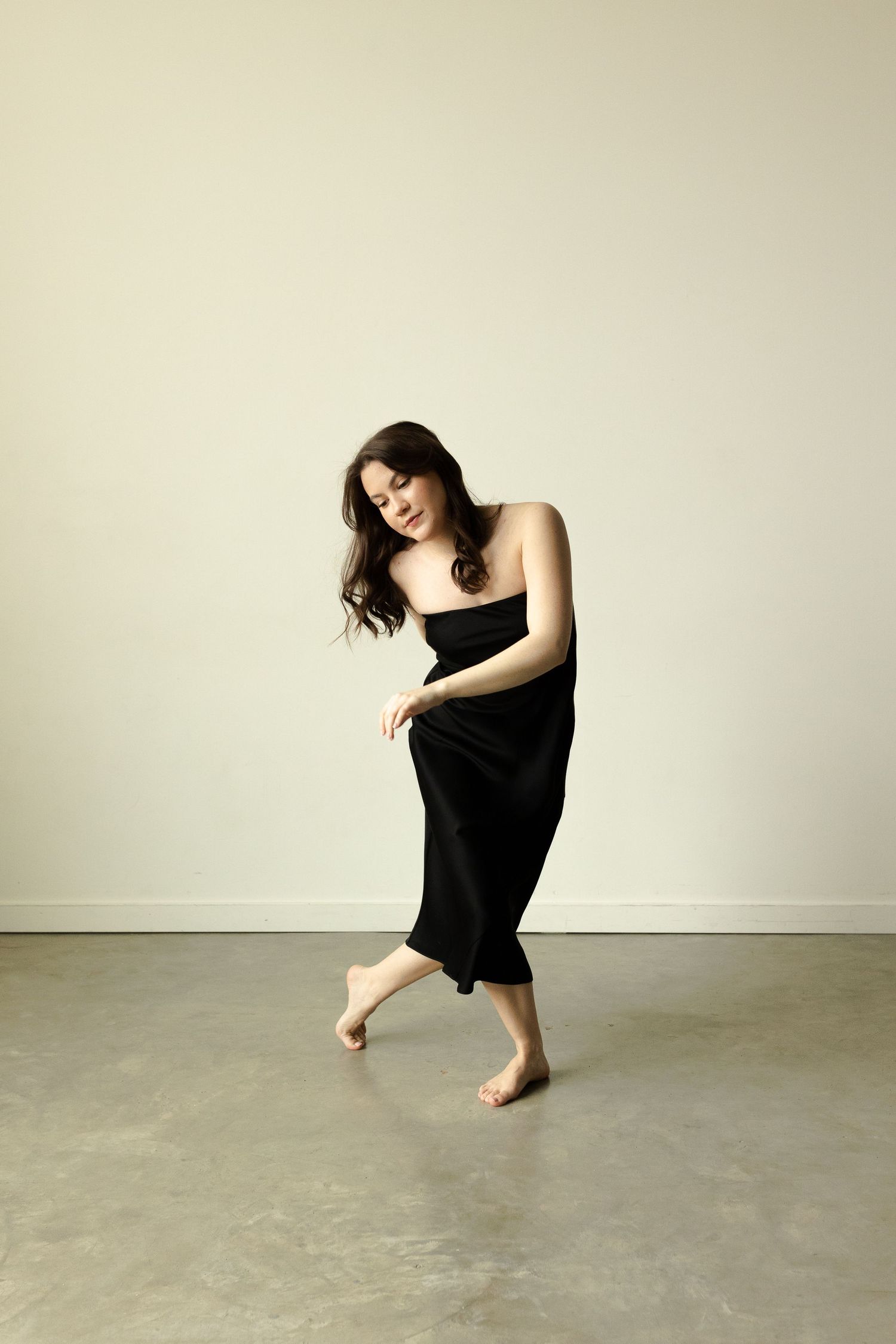 Artistic dance pose in black dress on concrete floor with natural lighting.