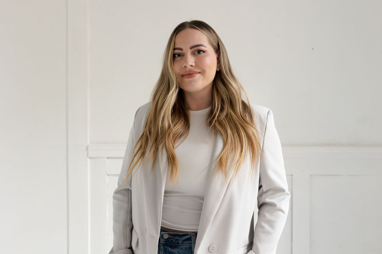 Fashion model wearing cream blazer and grey top in minimalist studio setting.
