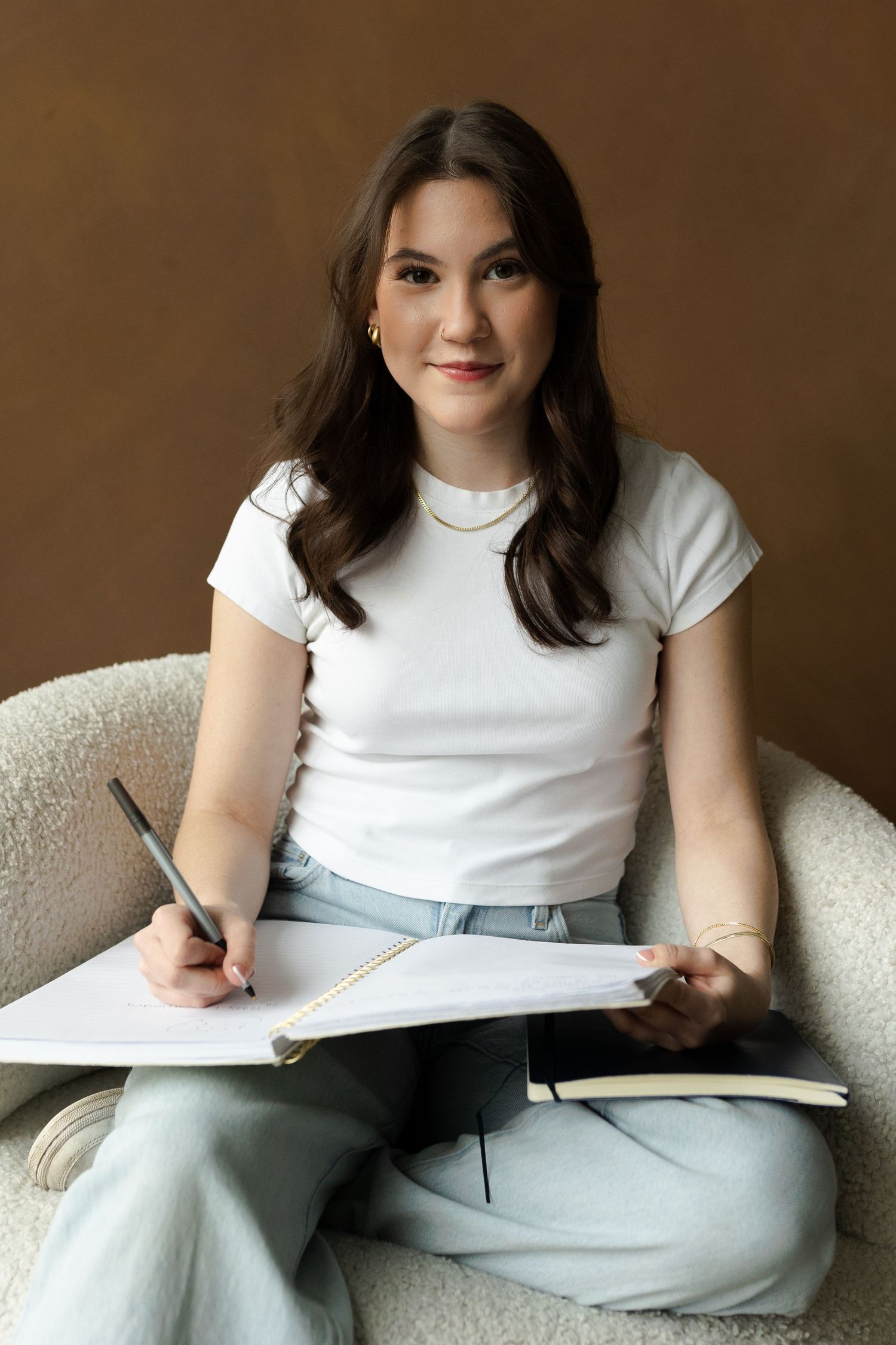 Person in white t-shirt and light pants writing in notebook while sitting on beige couch.