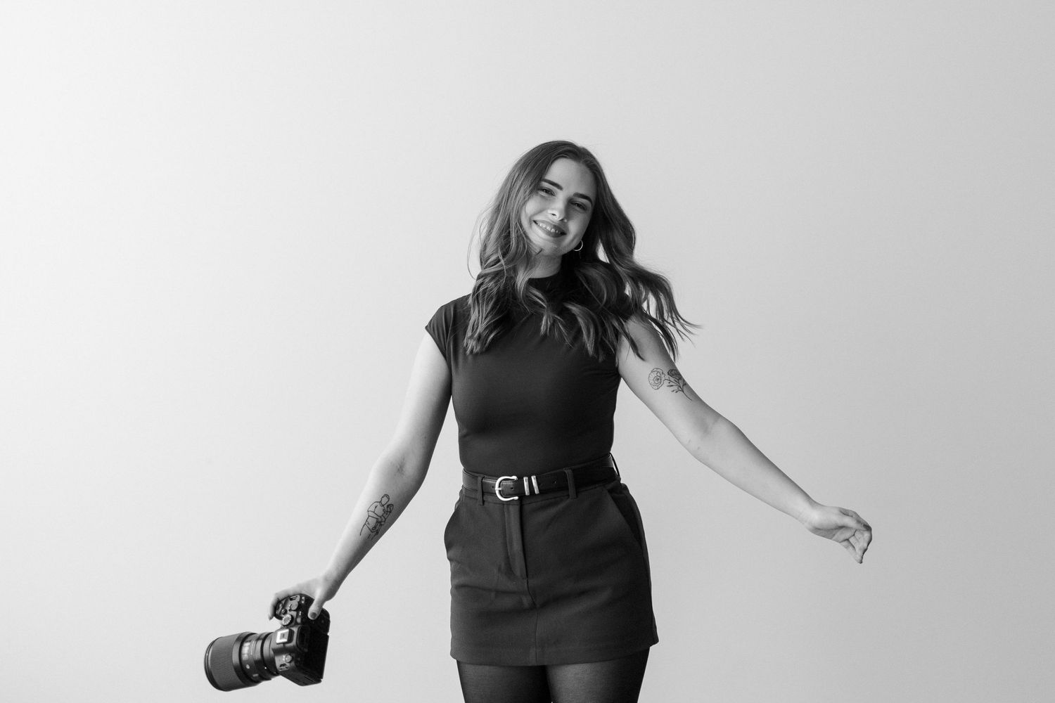 Black and white portrait of a person in a short dress holding a camera with windswept hair against light background.