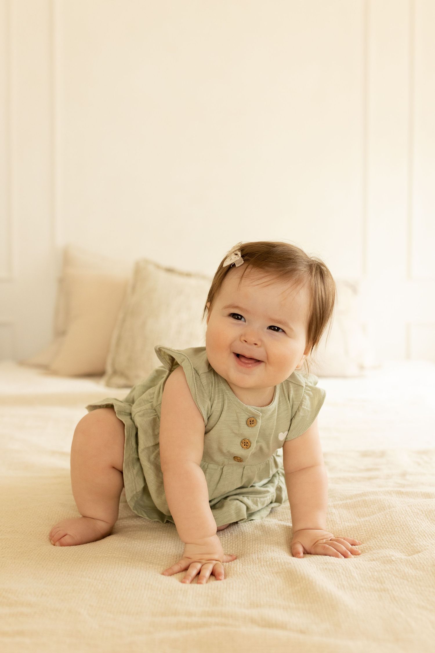 A sequence of four photos showing a baby in a sage green romper crawling and playing on a cream colored blanket.