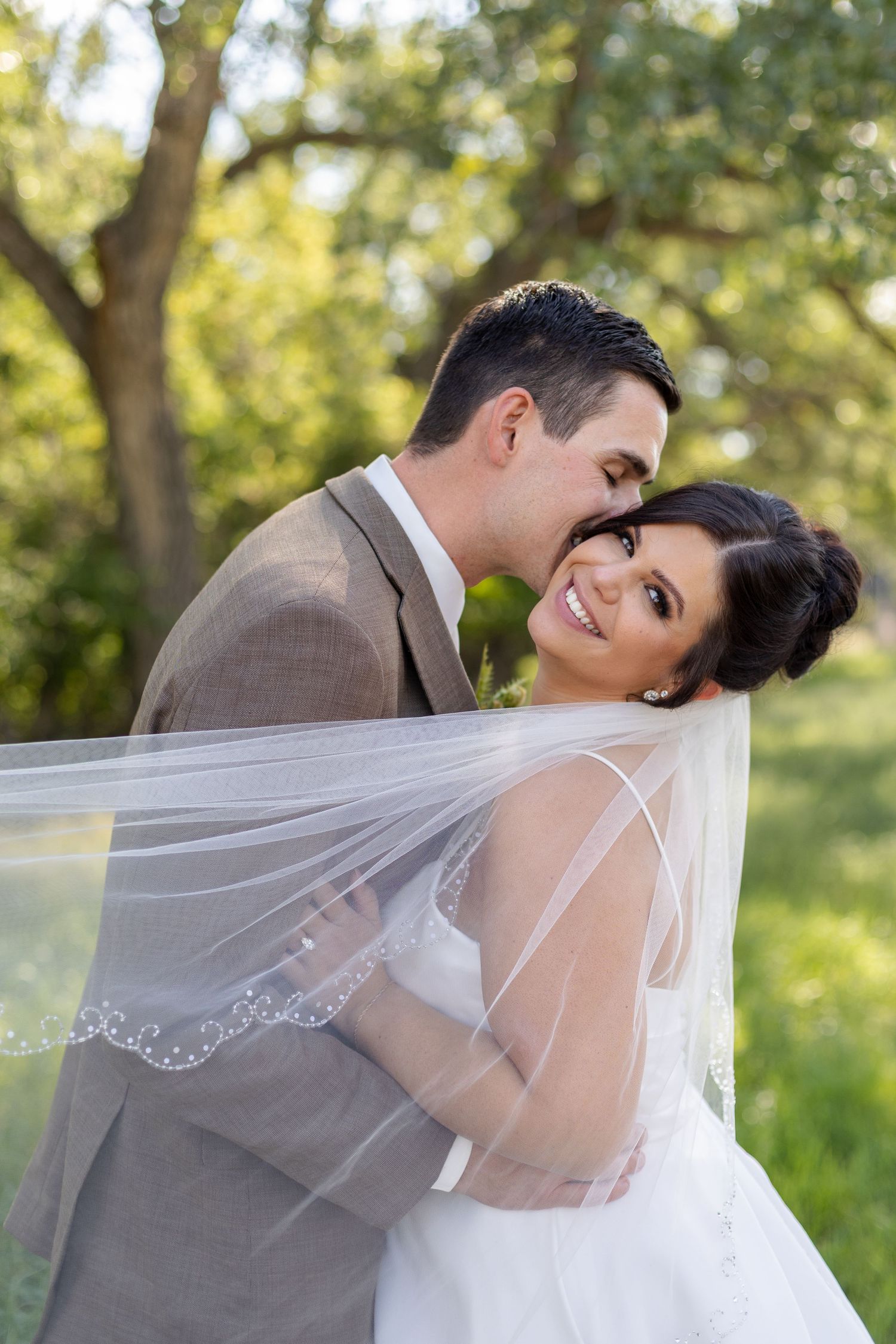 Tender wedding moments captured in a sunlit garden setting with flowing white veil and gray suit.