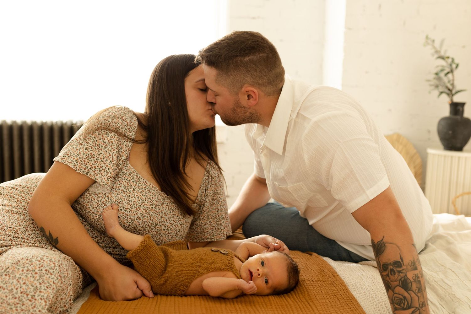 A couple shares an intimate moment while their baby lies between them on a bed in a sunlit room.