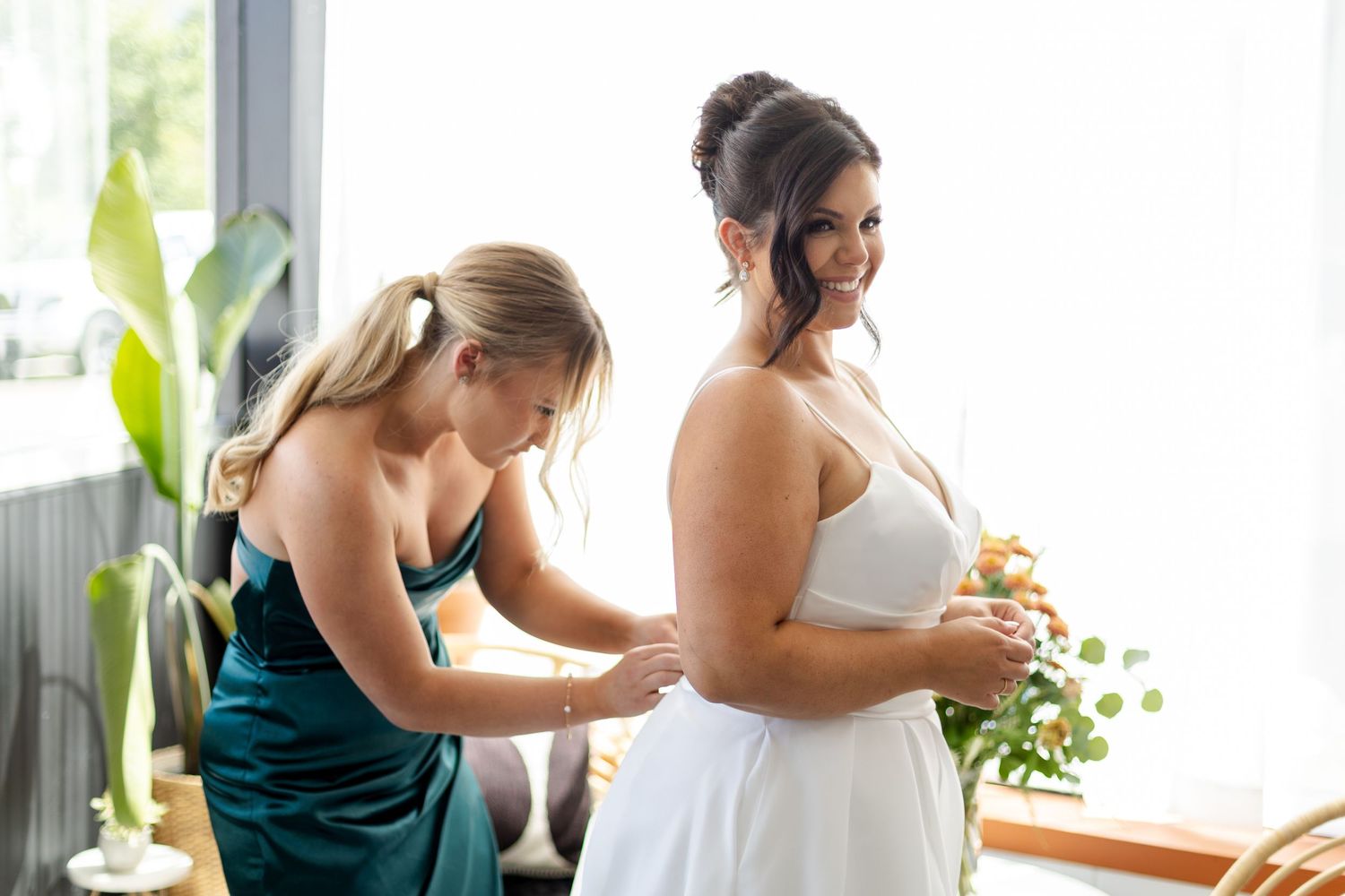 Wedding party members enjoy drinks and laughter during a pre-ceremony celebration in a bright sunlit room.