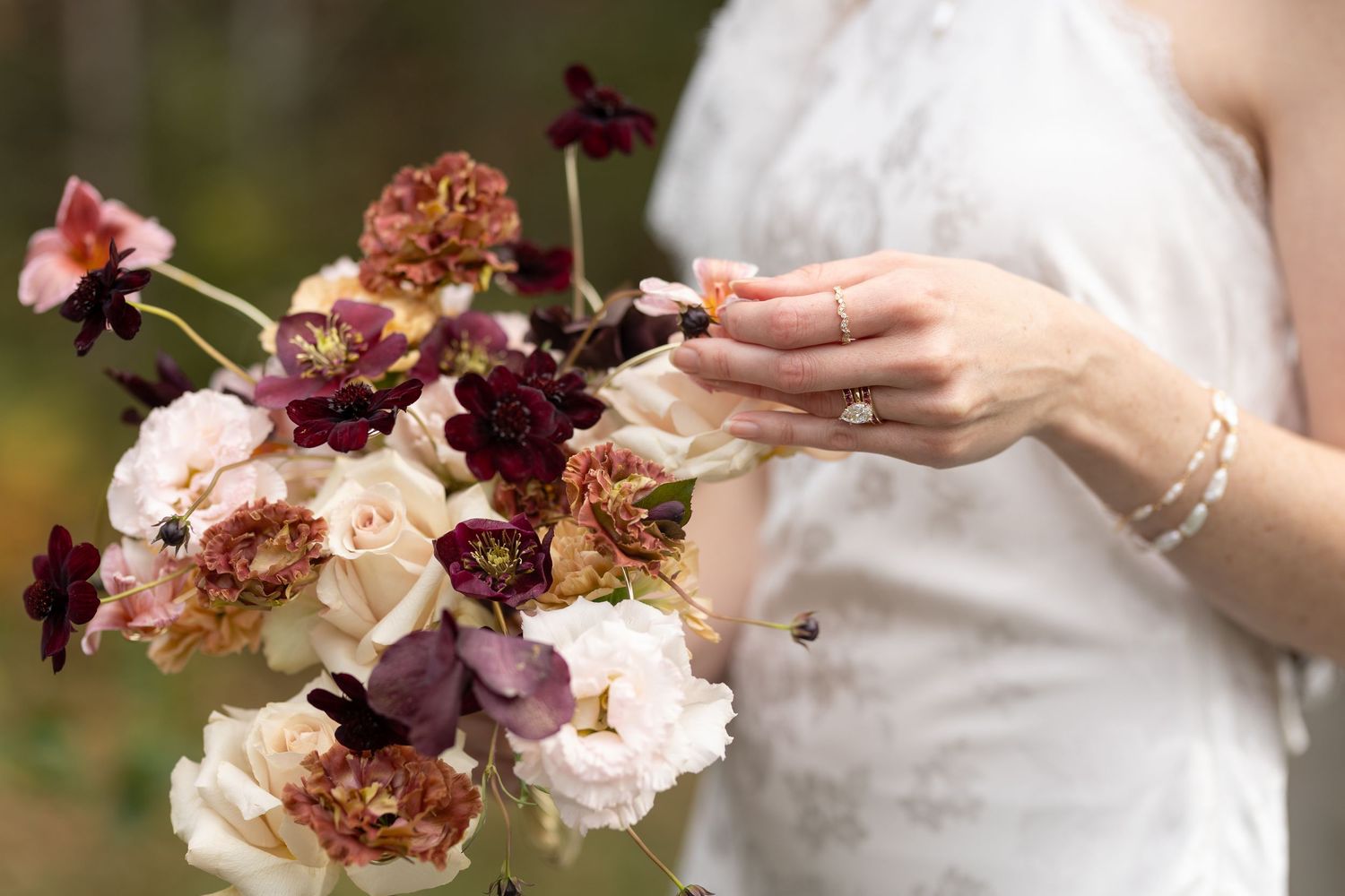 A hand wearing an engagement ring holds a romantic bouquet of burgundy and blush flowers against a blurred outdoor background.