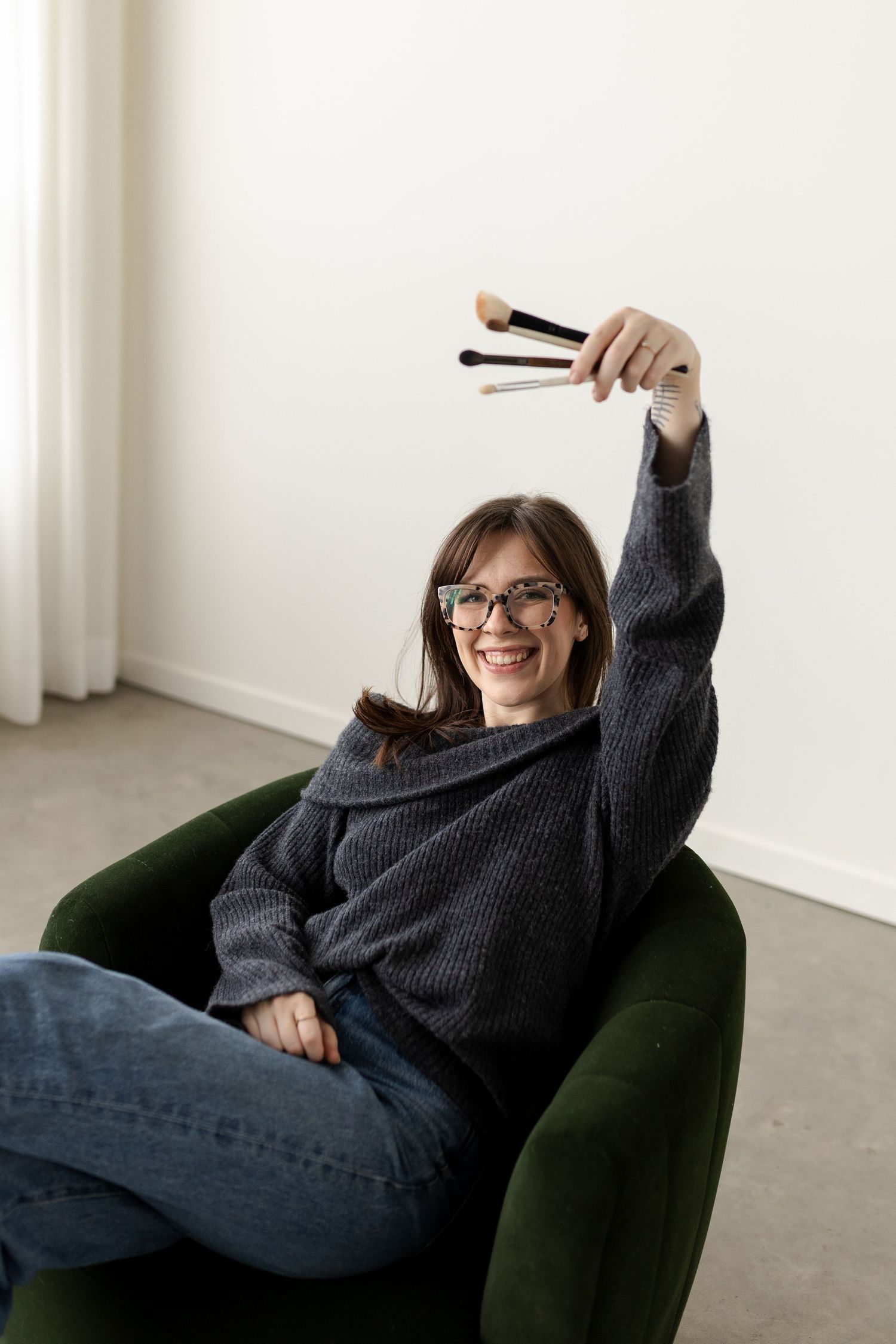 Person in glasses and gray sweater relaxes in green armchair while holding chopsticks up in playful pose.
