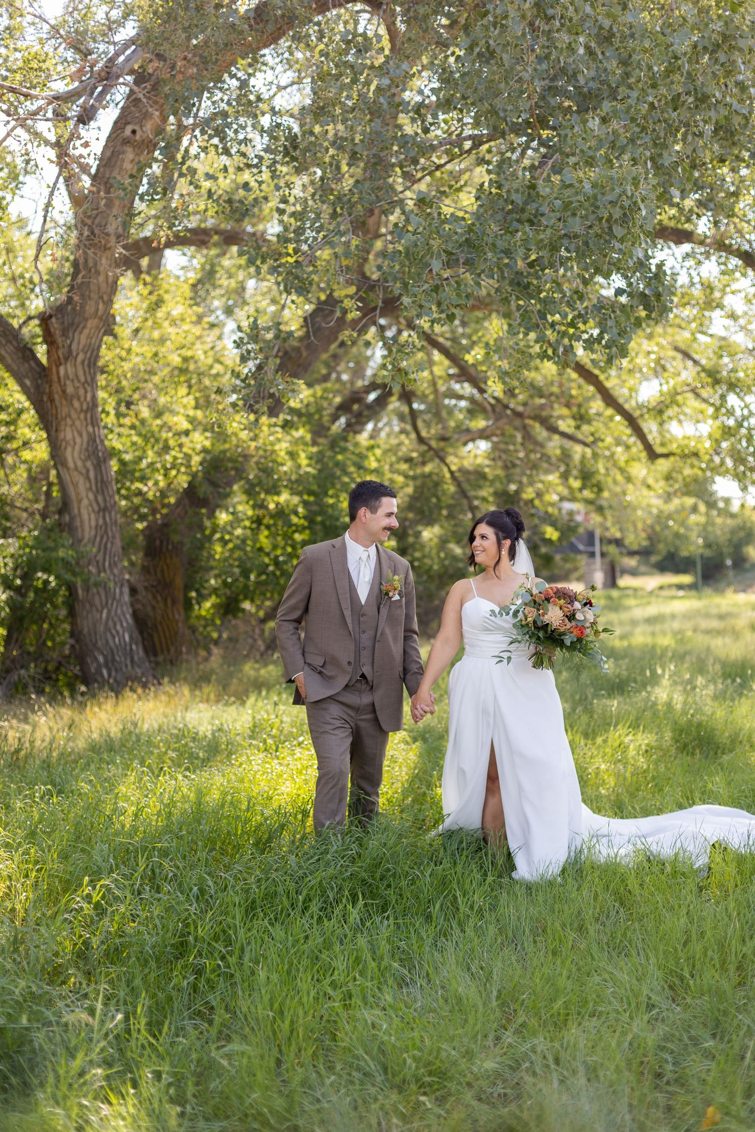 A couple in wedding attire walks together through a grassy meadow with trees in the background.