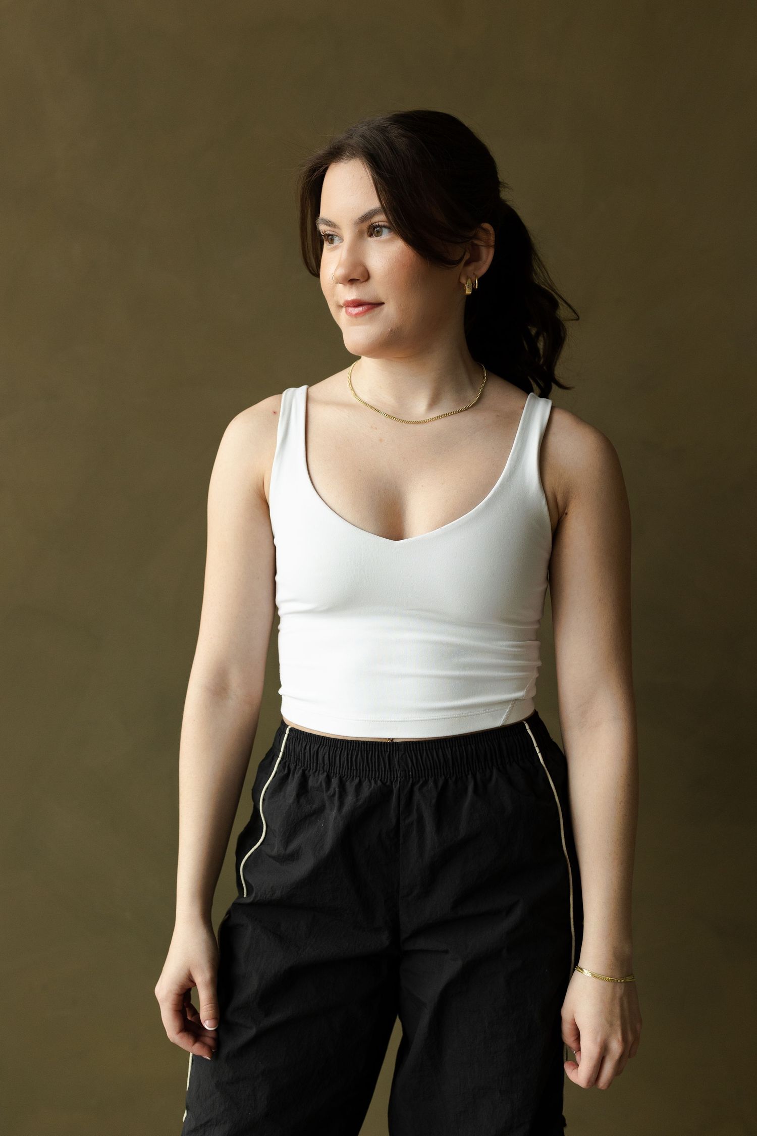 Woman in white tank top and black pants poses against olive green background in fashion photography series.