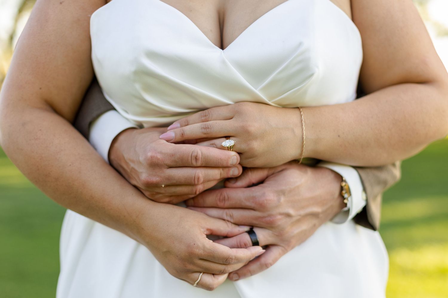 Close up view of hands with wedding rings embracing around a white wedding dress bodice in an outdoor setting.
