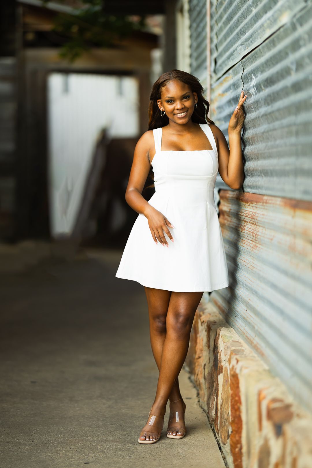 A person in a white summer dress poses against an industrial wall with metallic siding.
