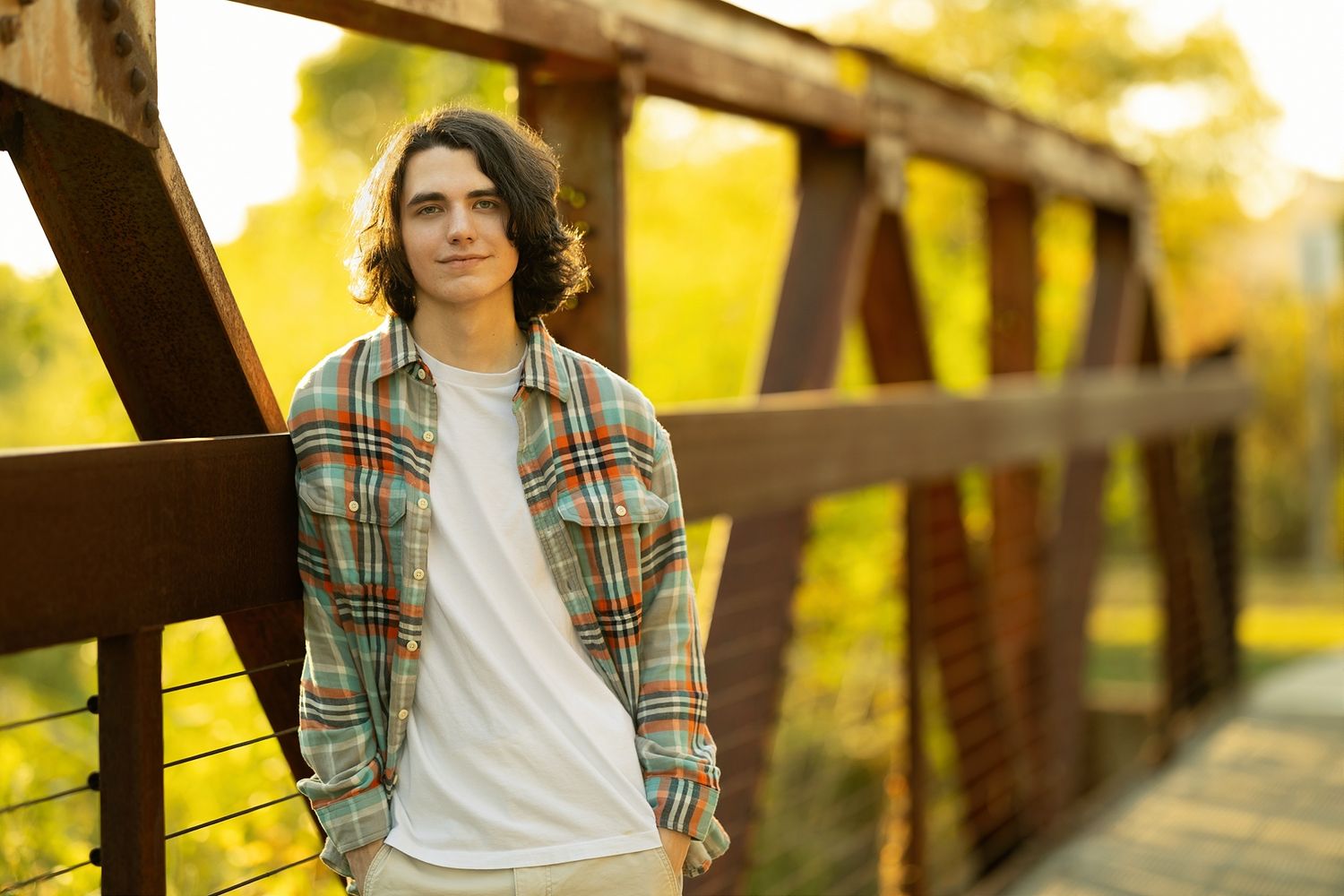 A person in a plaid shirt stands on a wooden bridge during golden hour with autumn trees in the background.