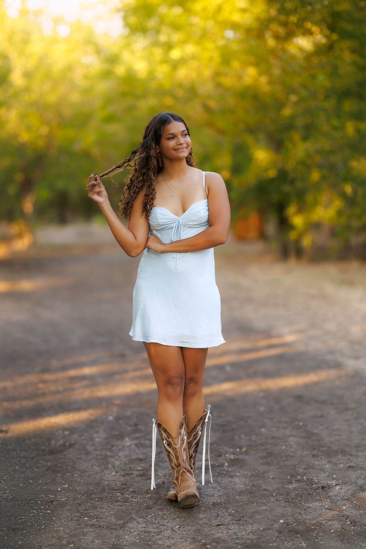 A woman in a white dress and cowboy boots stands on a dirt path with golden foliage behind her.