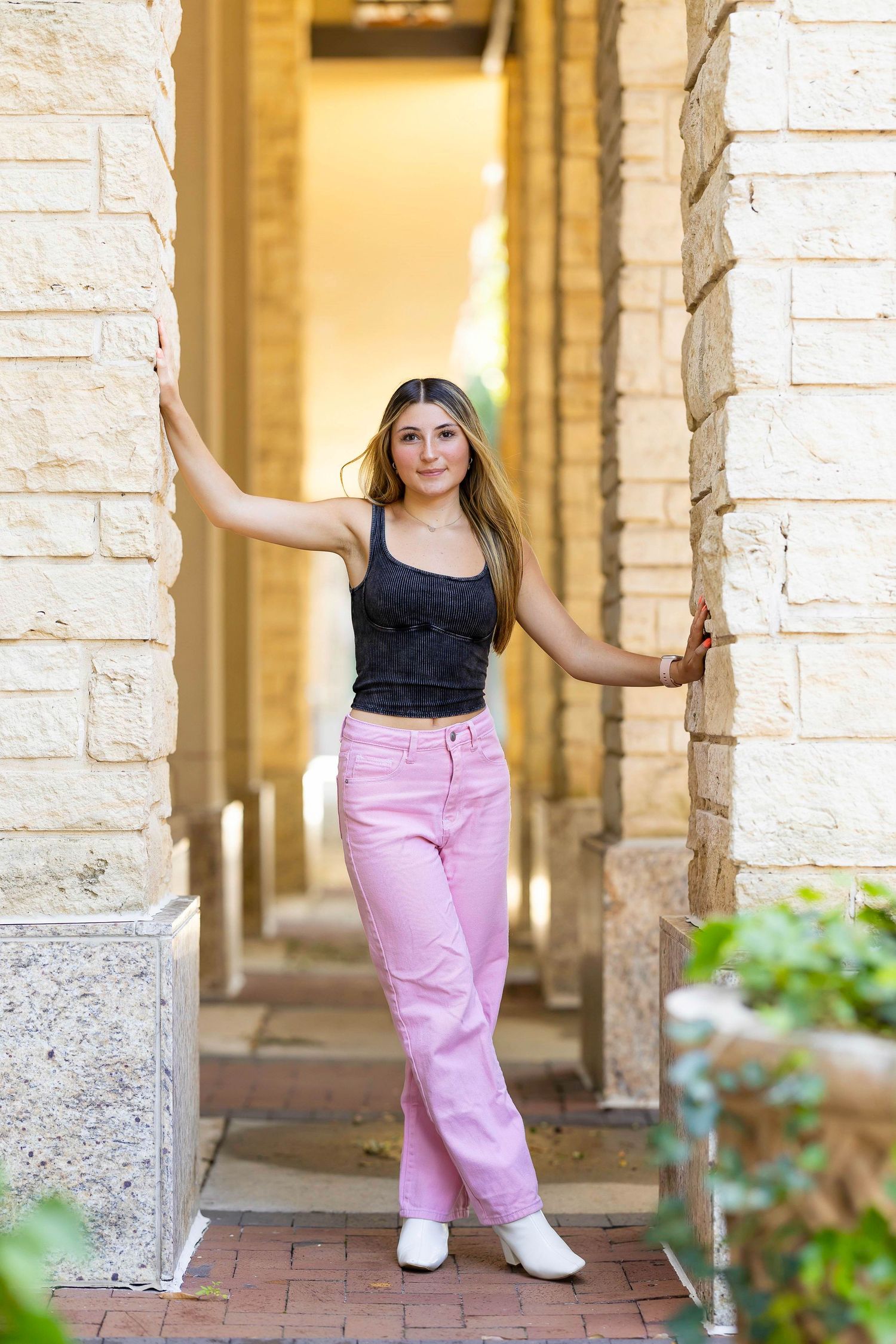 A person wearing a black top and pink pants poses between stone columns in an outdoor corridor.
